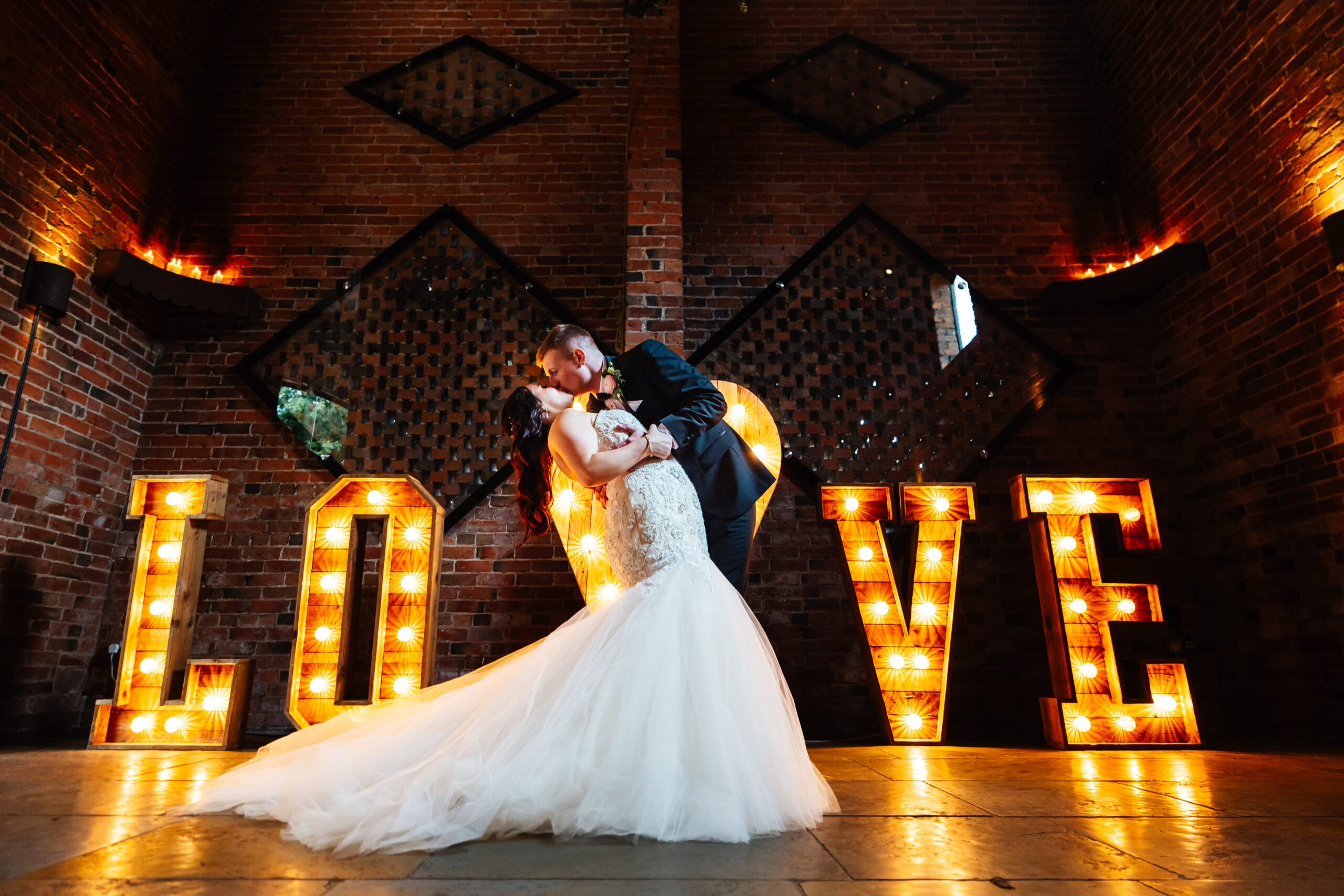 A photo of the bride and groom in front of light up letters that spell the word LOVE. The groom is holding his bride and tipping her back. They are holding hands and kissing.