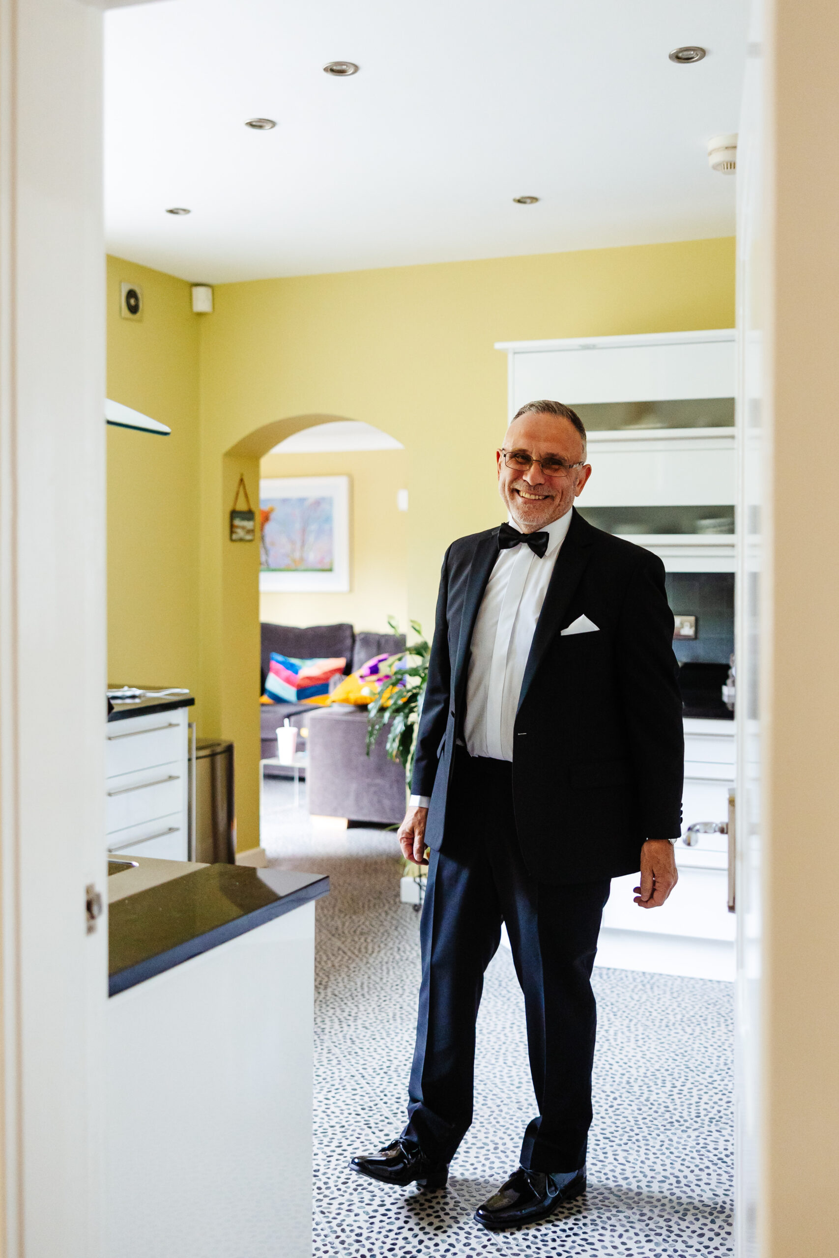 The father of the bride stood inside a kitchen. He has a smile on his face and is wearing a white shirt and black dinner jacket with a black bow tie.