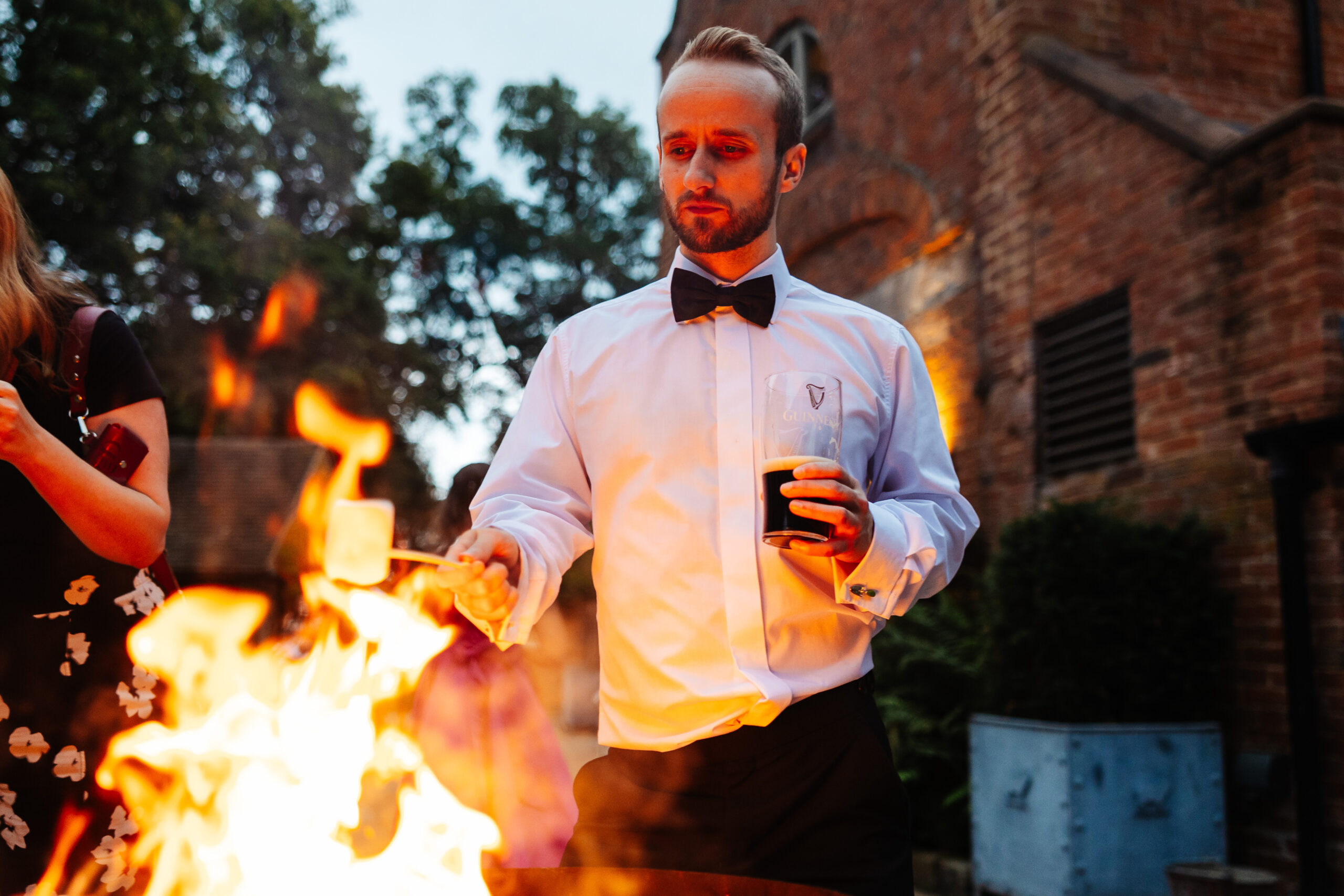 A man in a white shirt and black bow tie holding a marshmallow over an open fire. He has a pint of Guinness in his other hand.