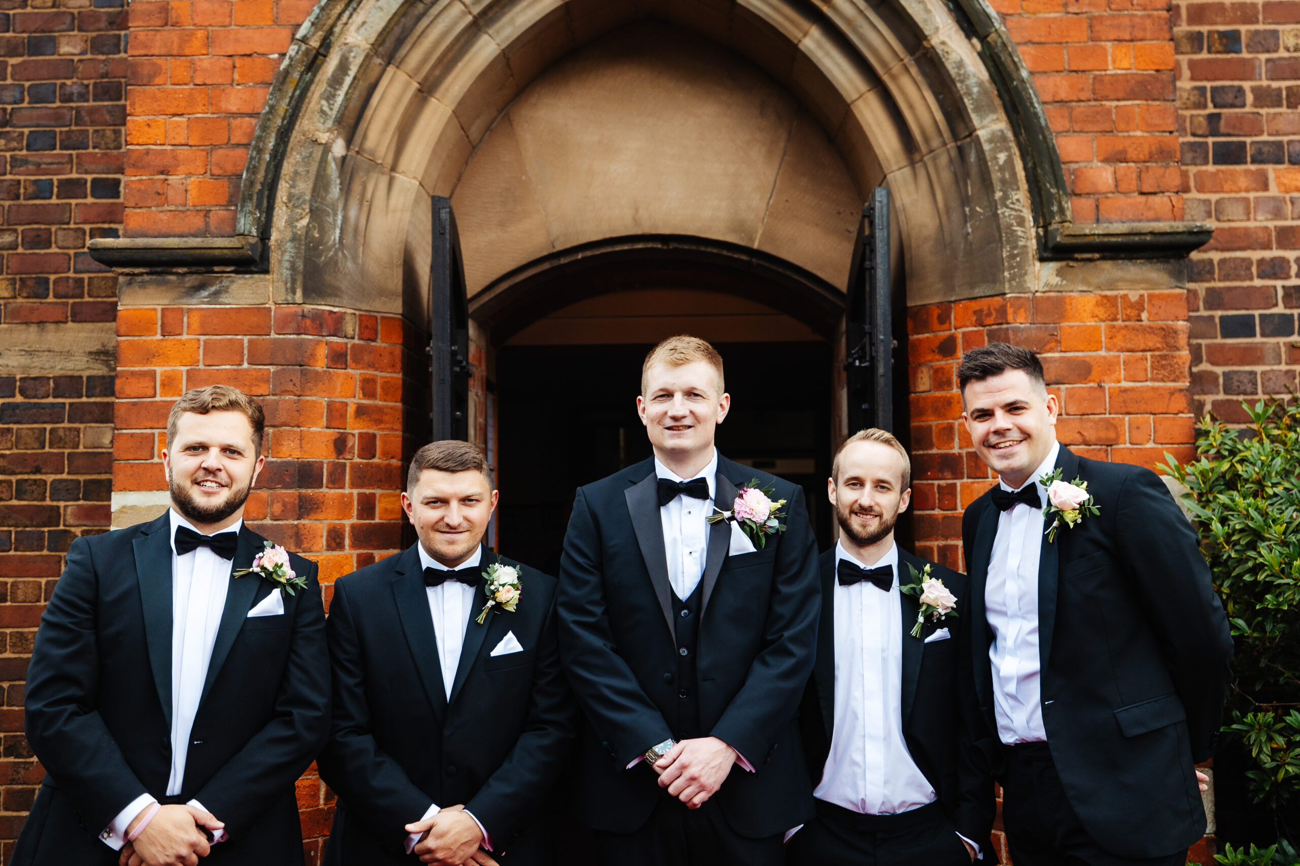 The groom and his groomsmen outside the church. They are all wearing black tie with black bow ties and are smiling.