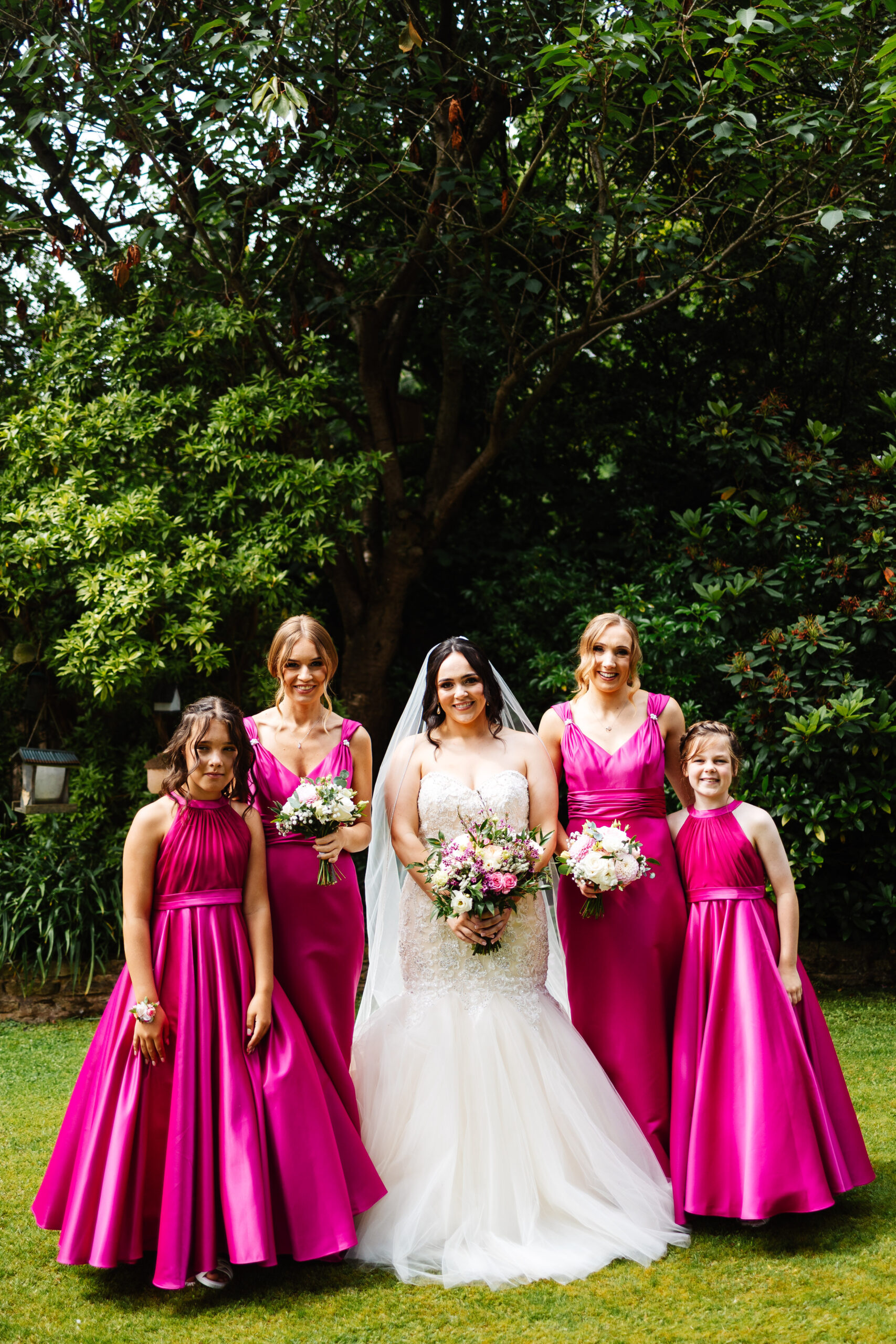 The bride and her bridal party in the garden. They are surrounding the bride and are all smiling. The bridal party are wearing hot pink dresses.