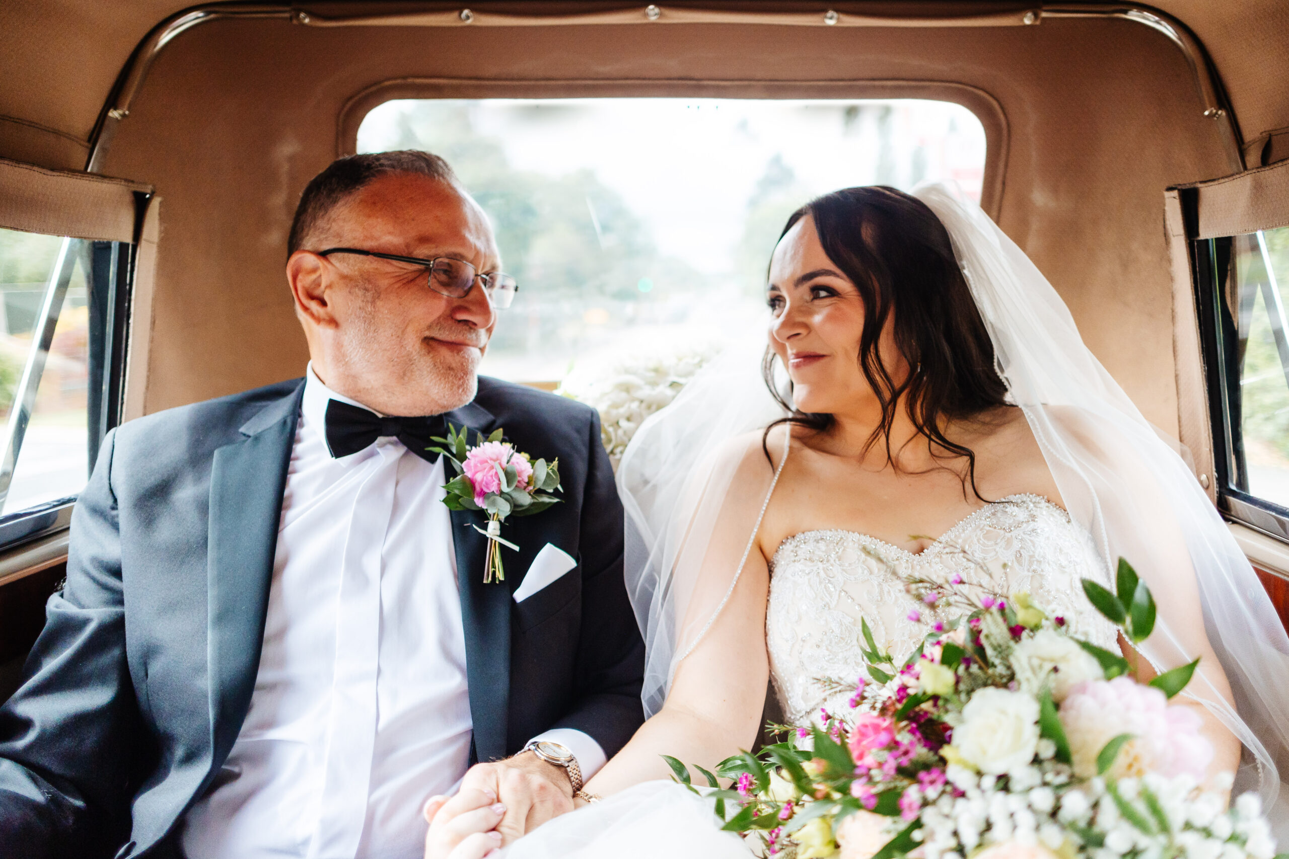 The bride and her father in the back of the car on the way to her wedding. They are looking at each other and smiling.