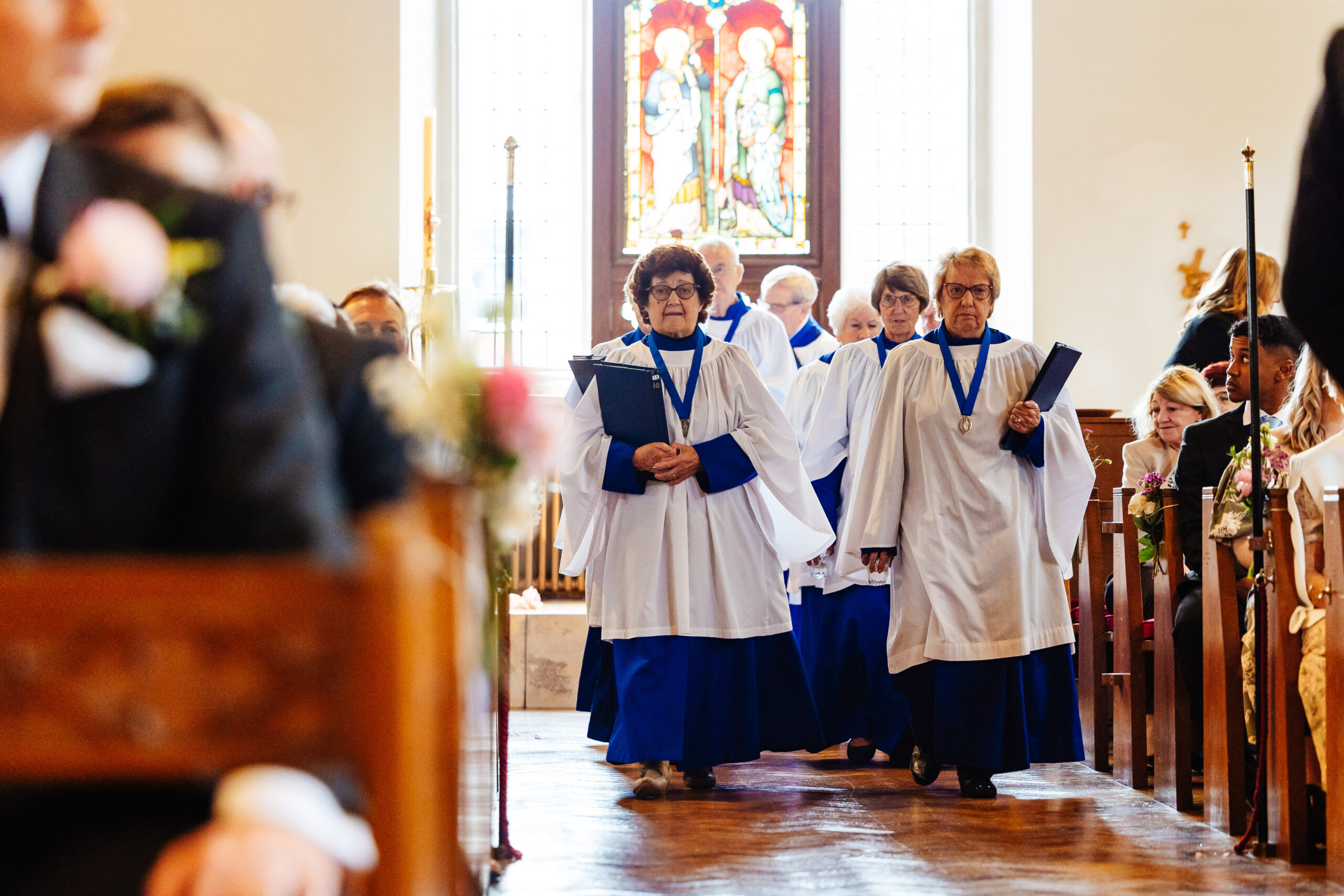 A choir walking in to the church. They are wearing white and blue tunics and are holding sheet music in their hands.