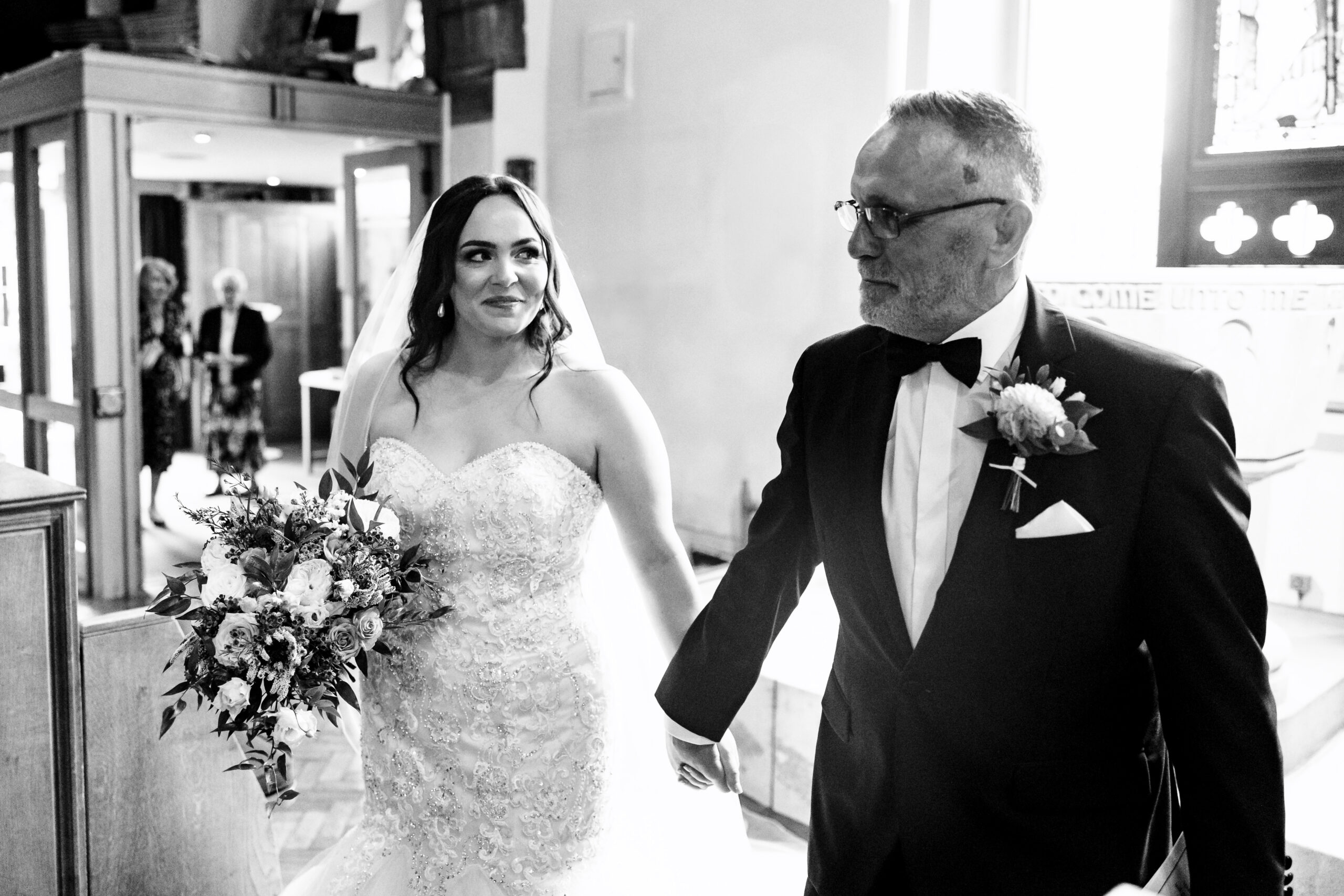 A black and white photo of the bride and her father before walking in to their wedding. The bride is looking at her dad and smiling.