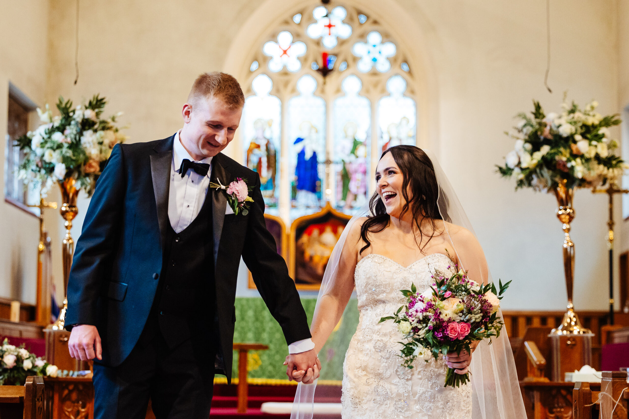 The bride and groom in the church just after they have got married. They are holding hands and the bride is looking at the groom and smiling.