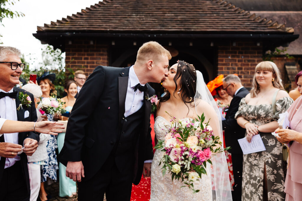 The bride and groom kissing outside surrounded by their friends and family. There is confetti in the bride's hair and in some of the guests' hands.