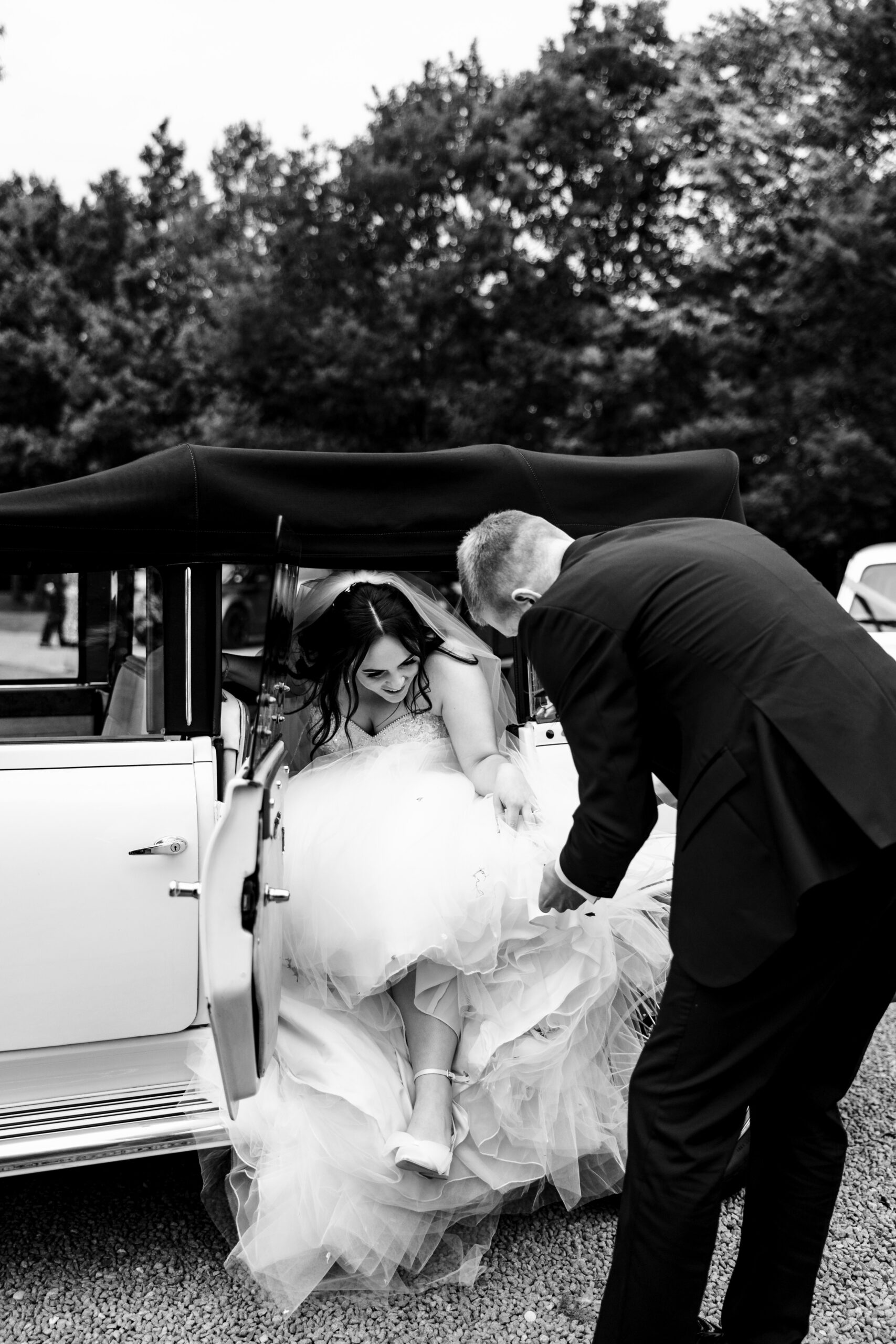 A black and white image of the bride getting out of the car. Her dress is very large and her groom is helping her get out of the car.