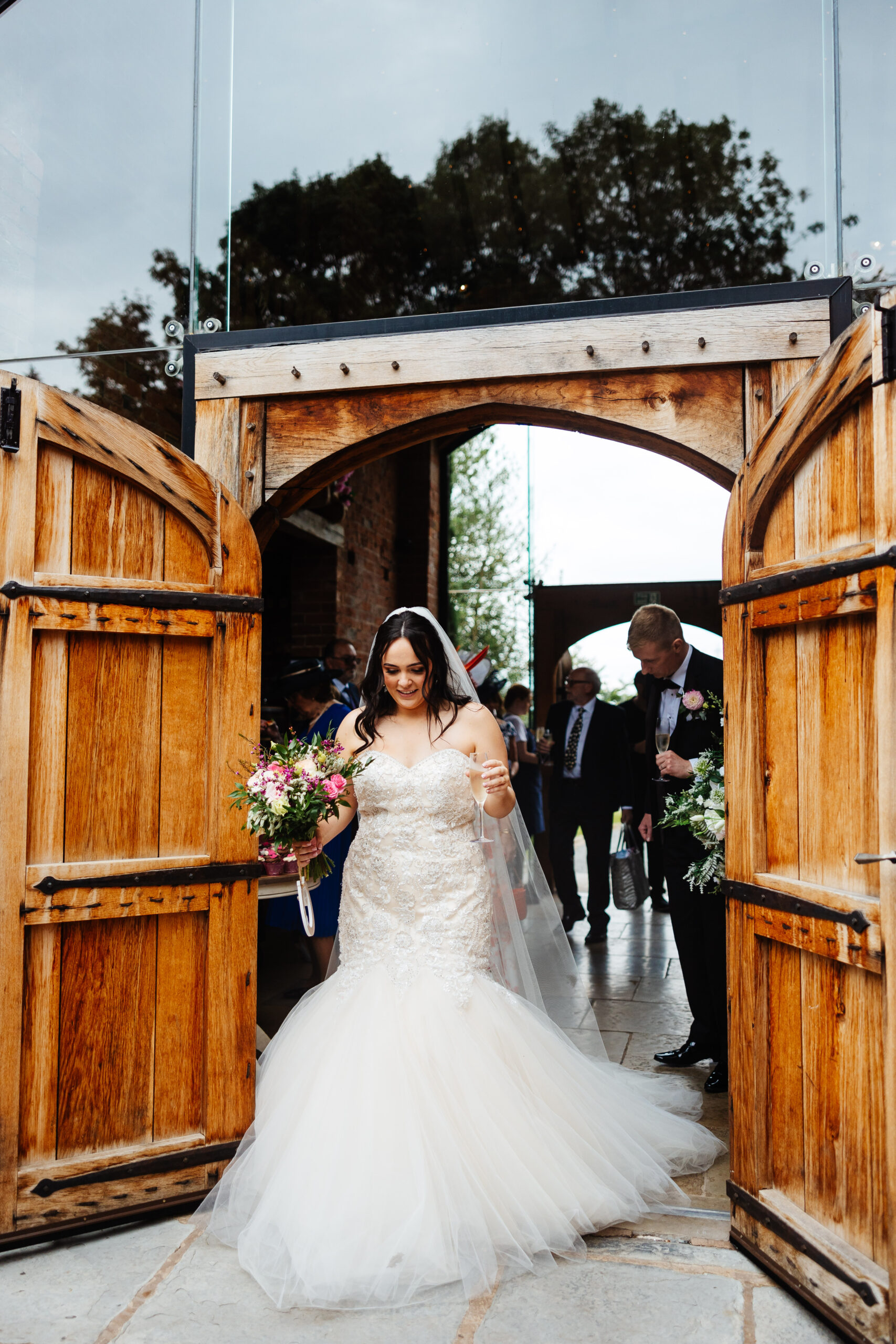 A photo of the bride walking out of two wooden, barn doors. She is holding flowers in one hand and a glass of champagne in the other. Her dress has a poofed bottom and is trailing on the floor behind her.