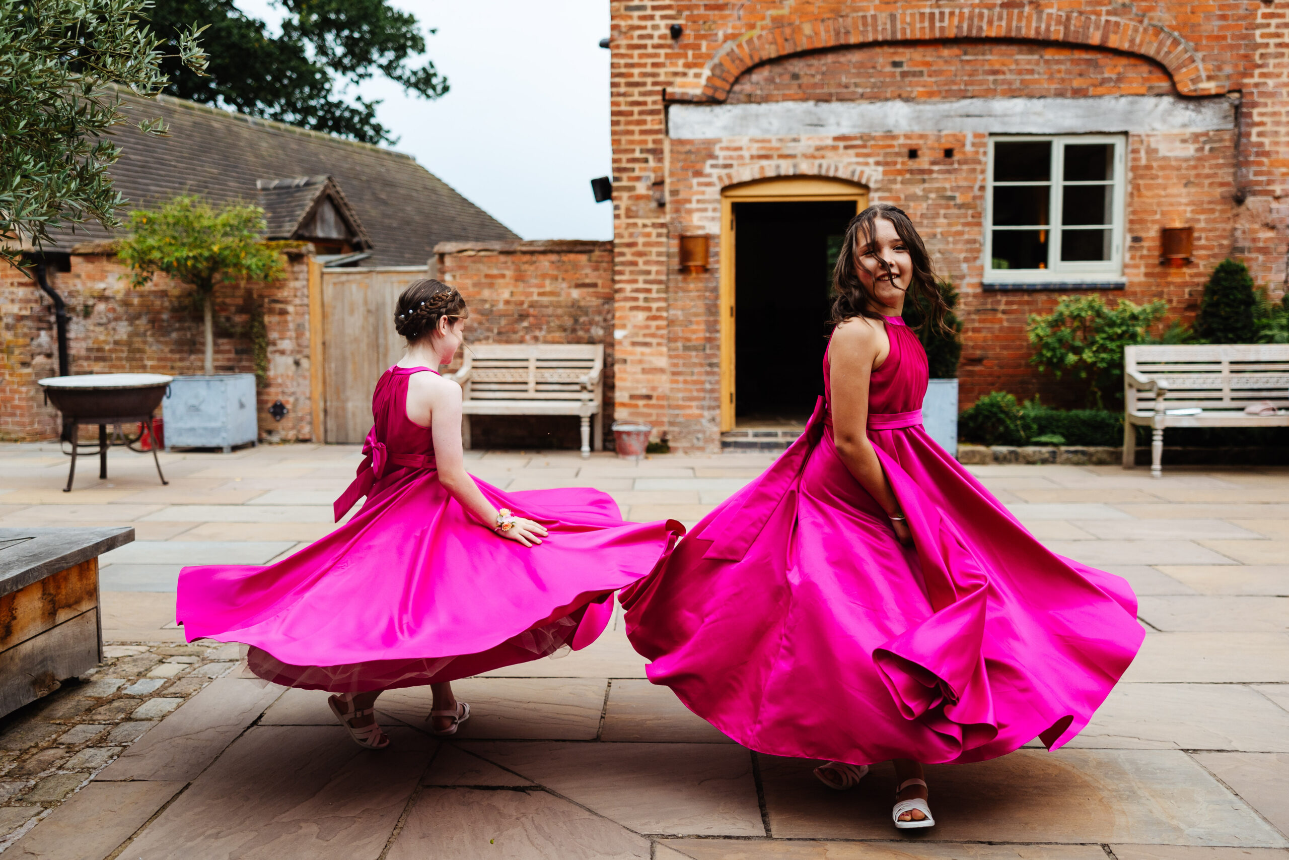 Two of the bridesmaids outside. They are wearing bright pink dresses and are twirling them in the air so the bottom of the dress are floating in the air.