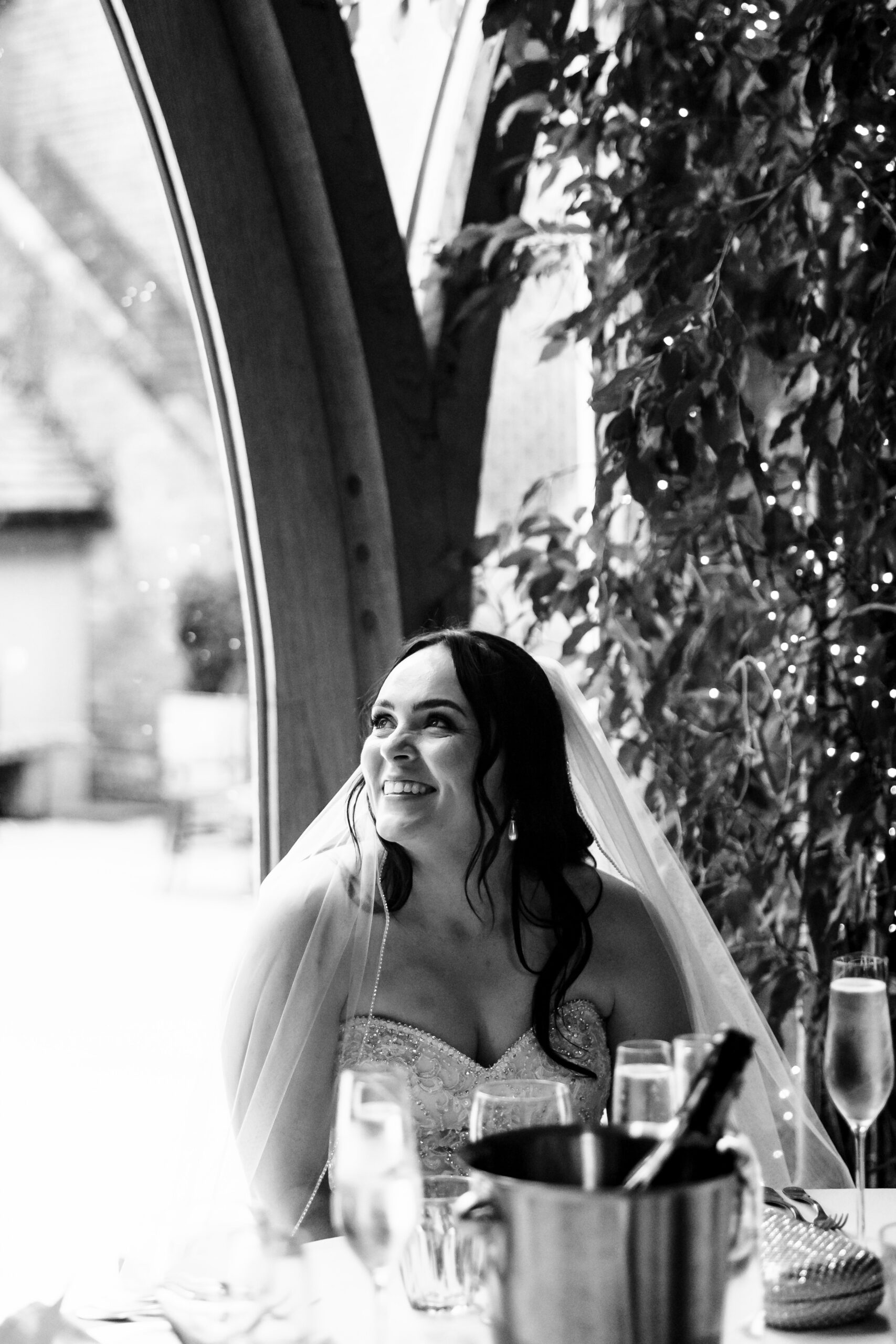 A black and white image of the bride looking up at her groom during his speech. She is smiling.