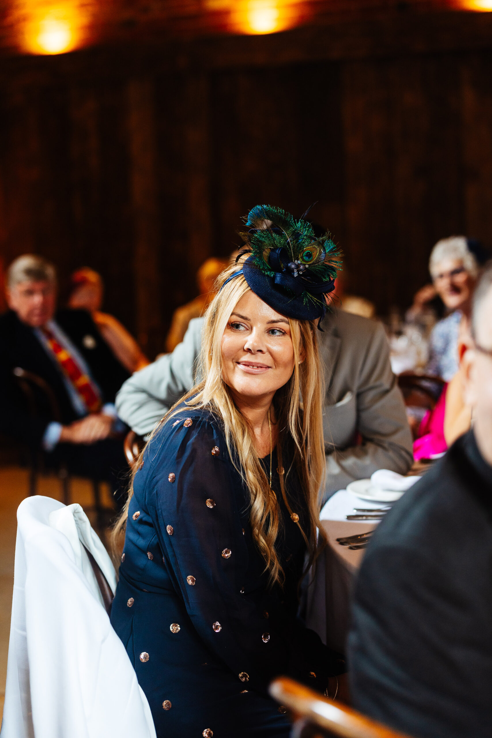 A guest in a blue dress and matching hat. She is looking at the bride and groom during speeches and is smiling.