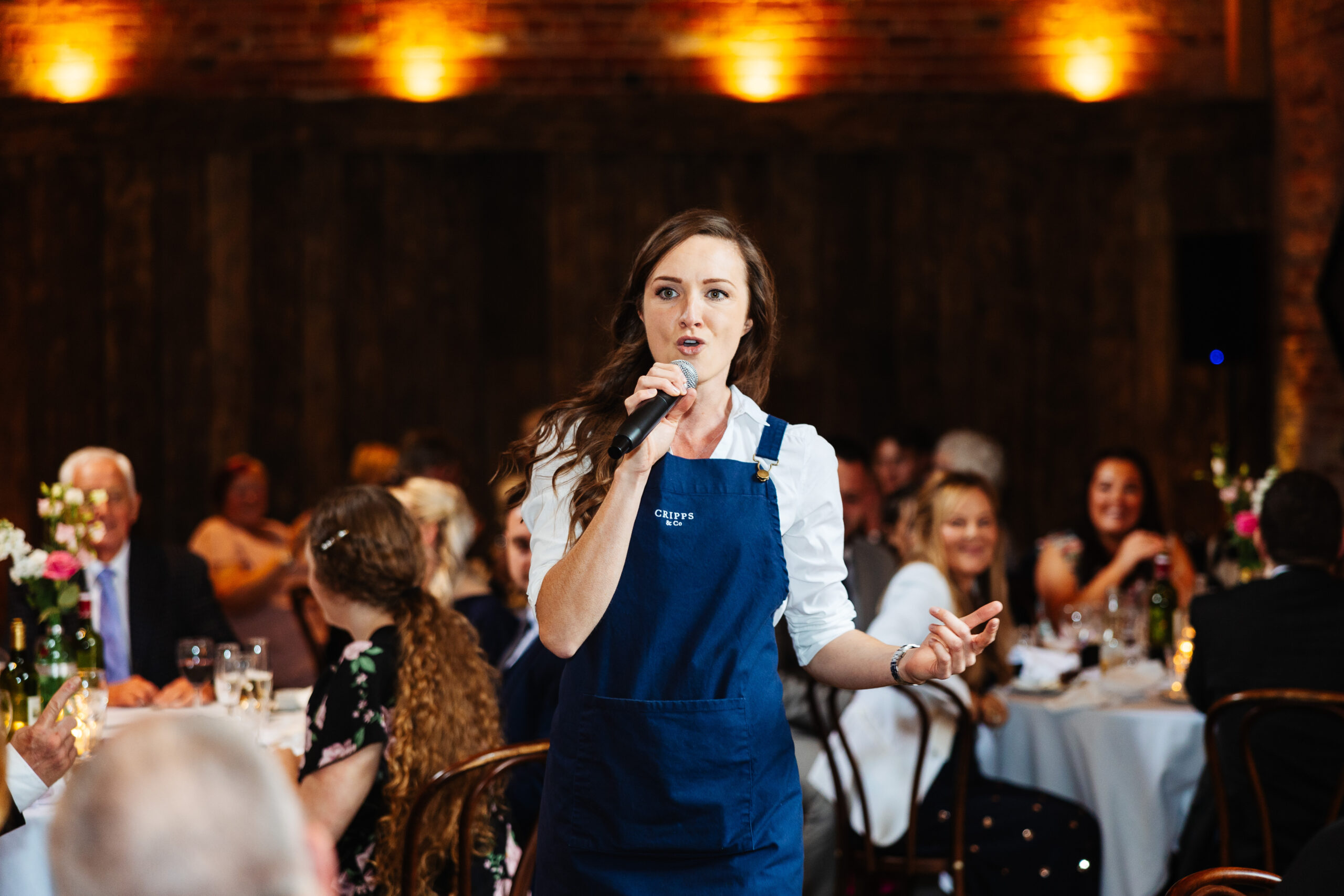 A waitress in a white shirt and a blue pinny. She is singing as a surprise and is holding a microphone.