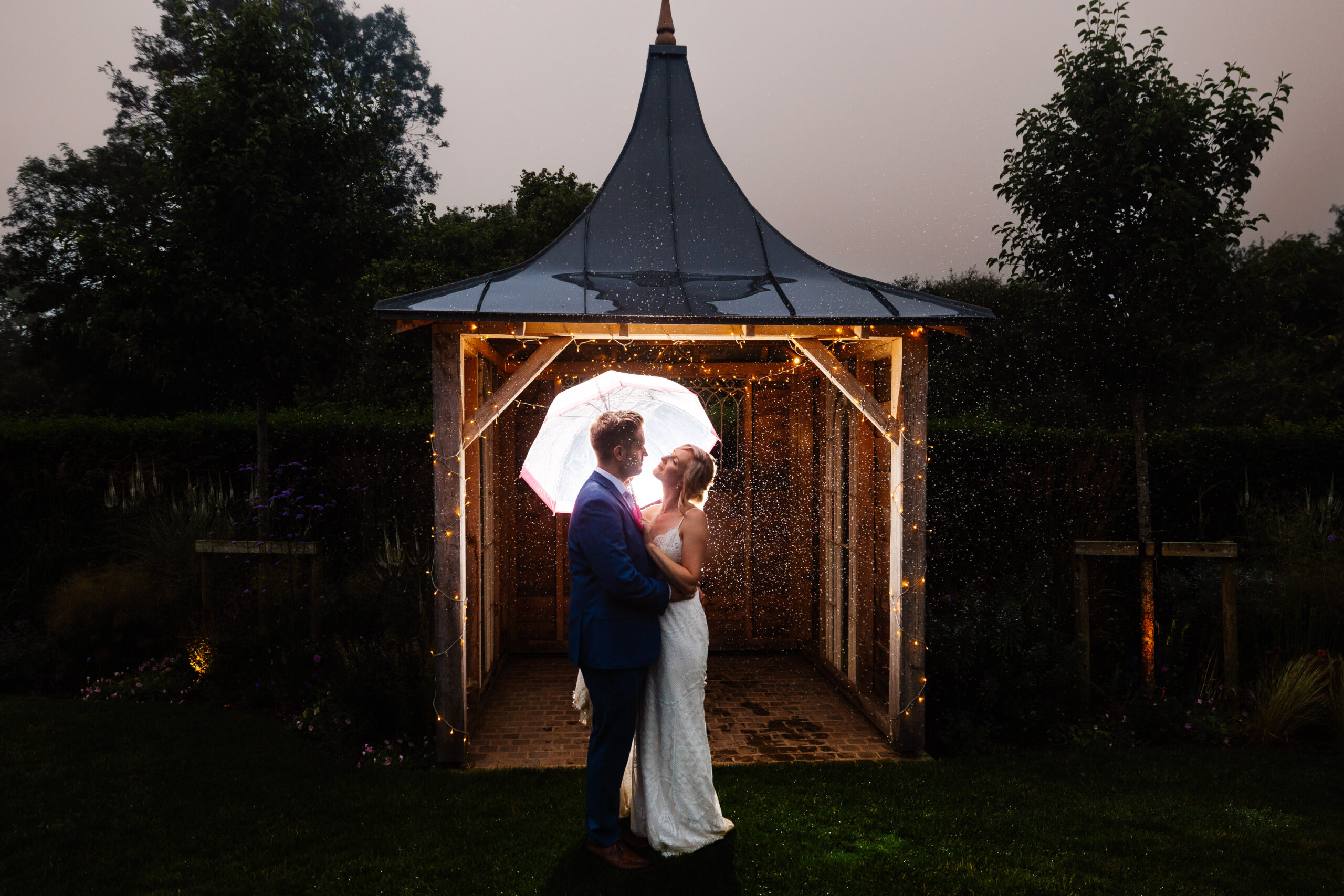 The bride and groom outside. They are holding an umbrella as it is raining. There are fairylight around them and they are looking lovingly at one another.