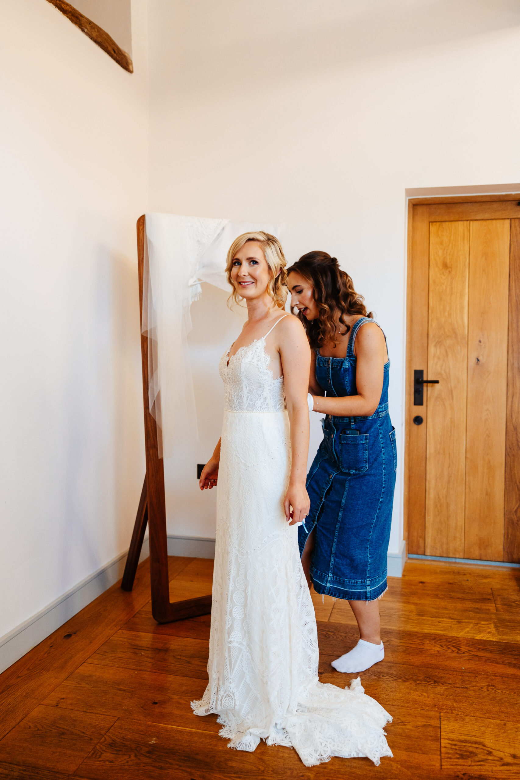 The bride in her wedding dress. She has a long wedding dress on with thin straps. One of her bridesmaids is helping her in to it. The bride is looking at the camera and smiling.