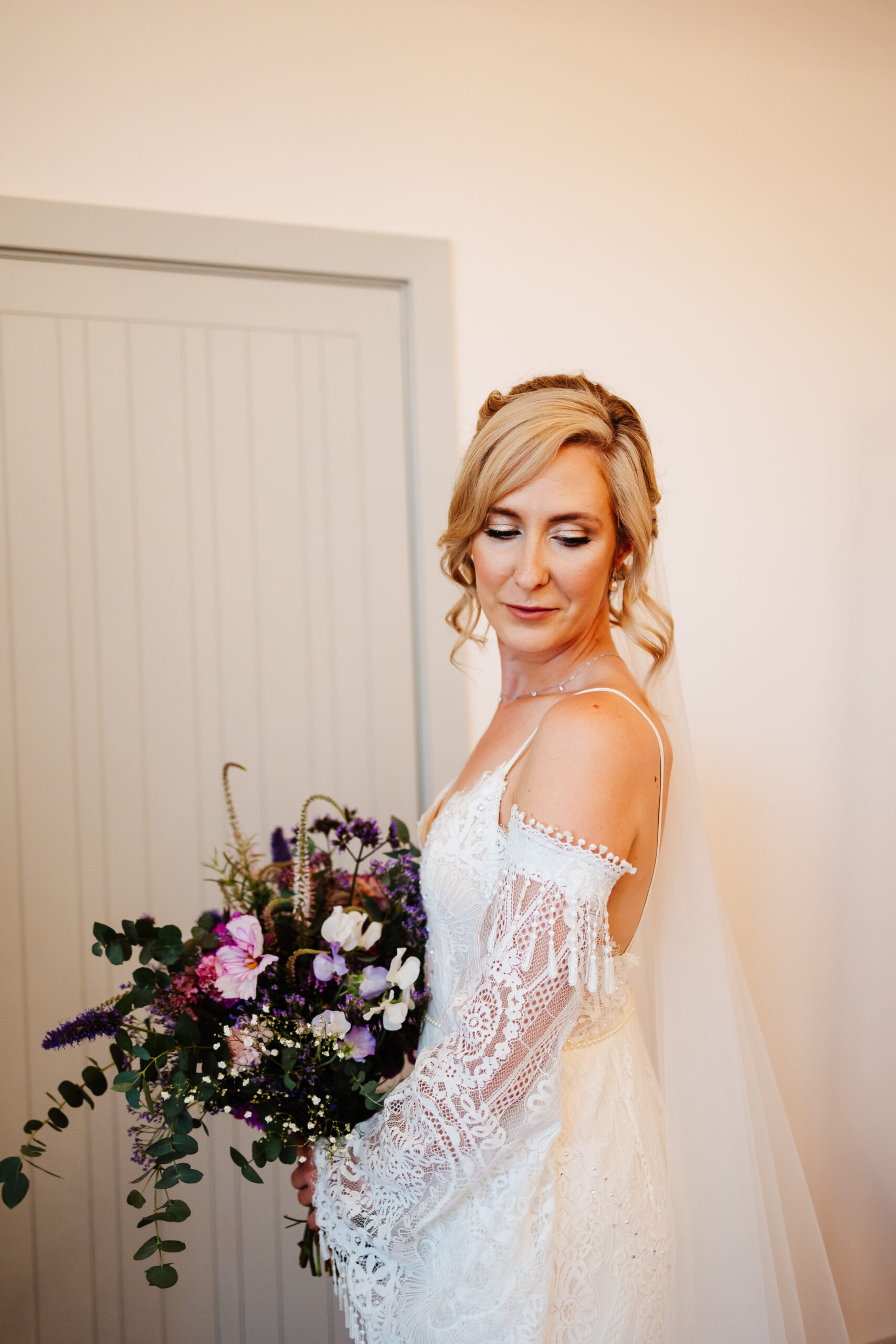 A photo of the bride. She is in her wedding dress which has laced detailed, off the shoulder sleeves. She is holding her bouquet of flowers which are tones of purples and pinks.