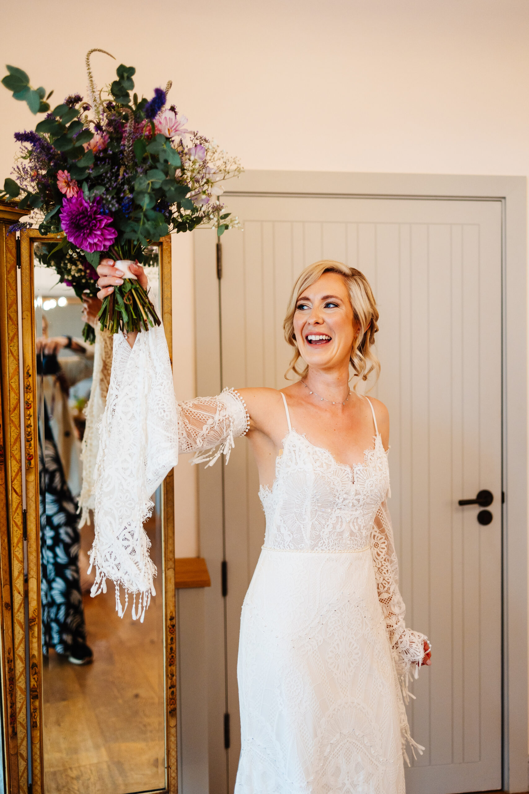 The bride lifting up her bouquet. She is smiling at her bridesmaids off screen. Her lace sleeve is hanging down. 