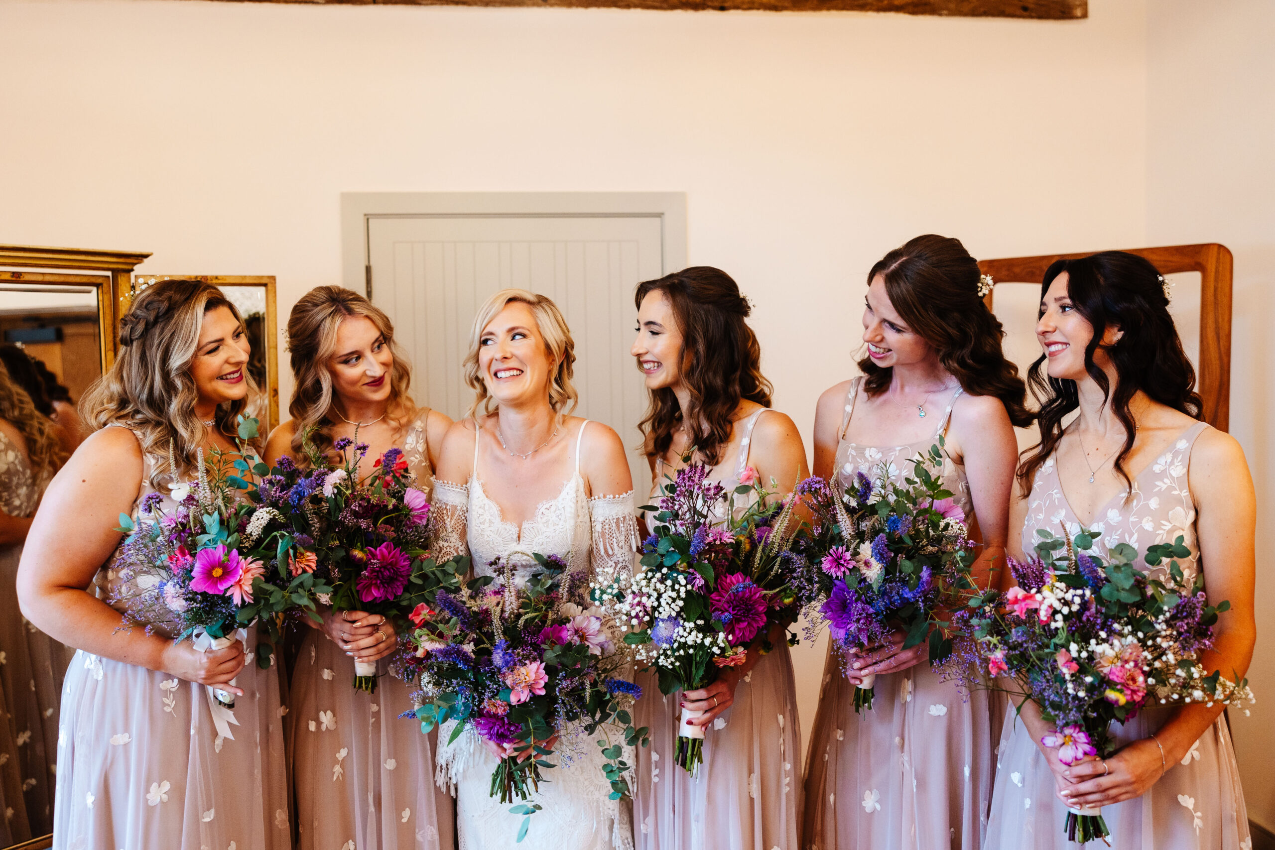 The bride and her bridesmaids. They are looking at the bride and the bride is looking at one of them. The bridesmaids are in pale pink dresses with white lace flowers on.