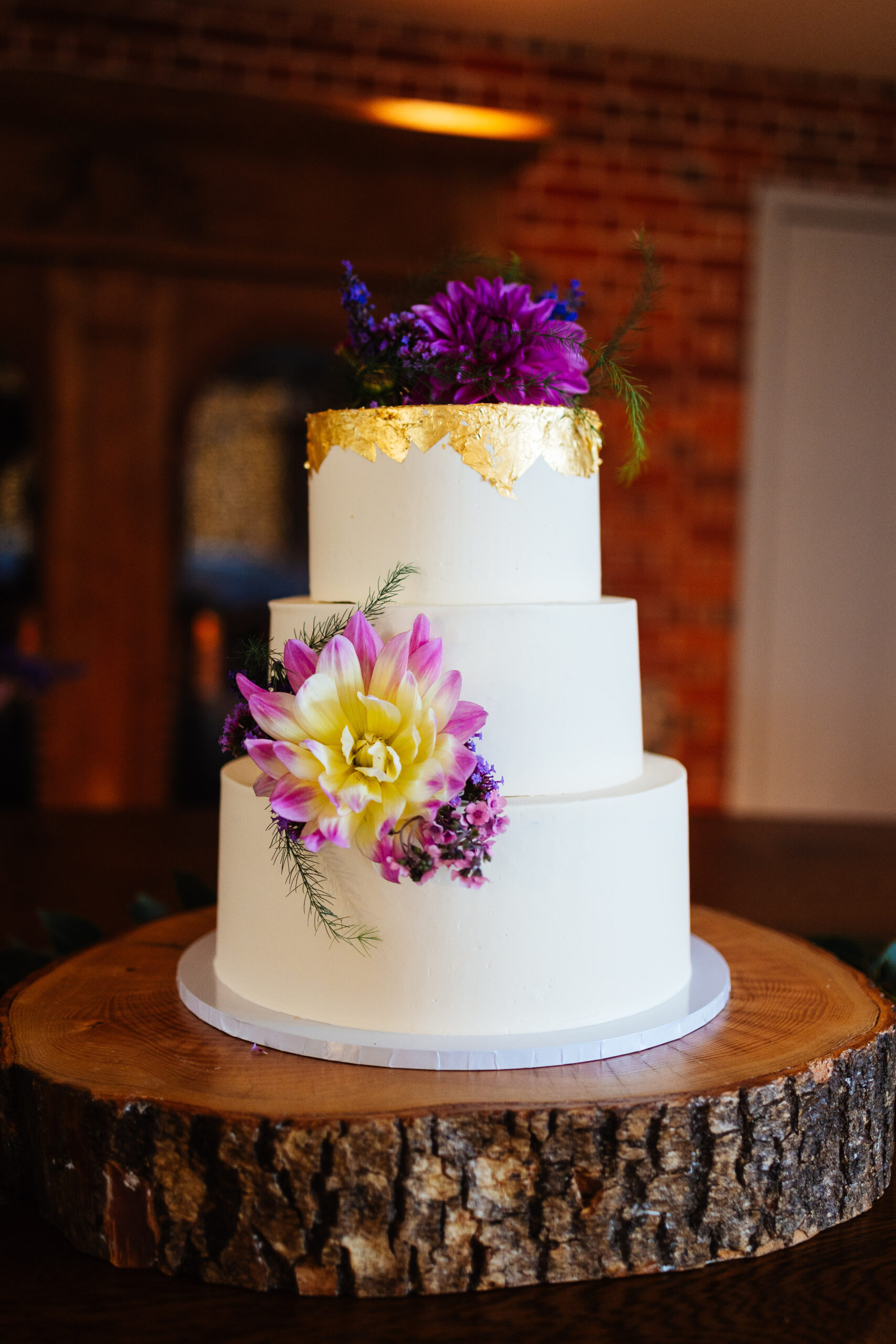 A three tiered, white cake. The top layer has gold foil detail at the very tip of the cake and has purple flowers laid on top.
There is also a pink and yellow flower placed neatly between the second and third layer.