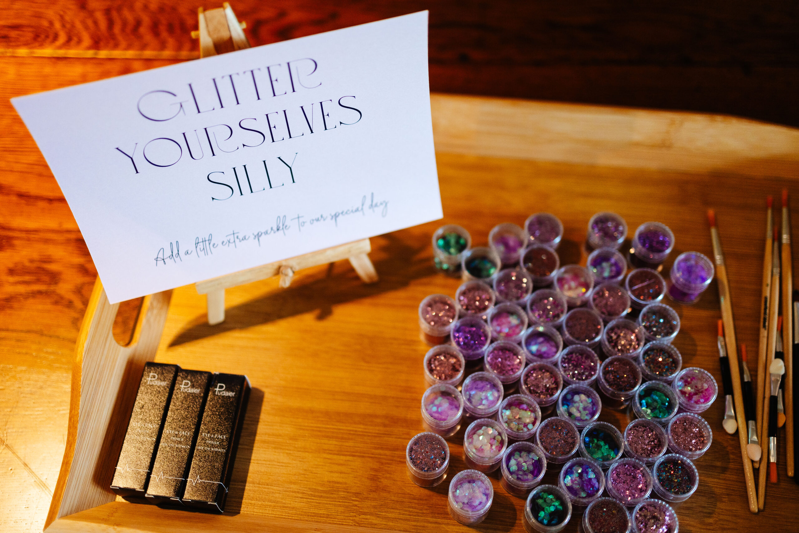 Tubs of glitter on a table with paint brushes. The sign says 'glitter yourself silly'