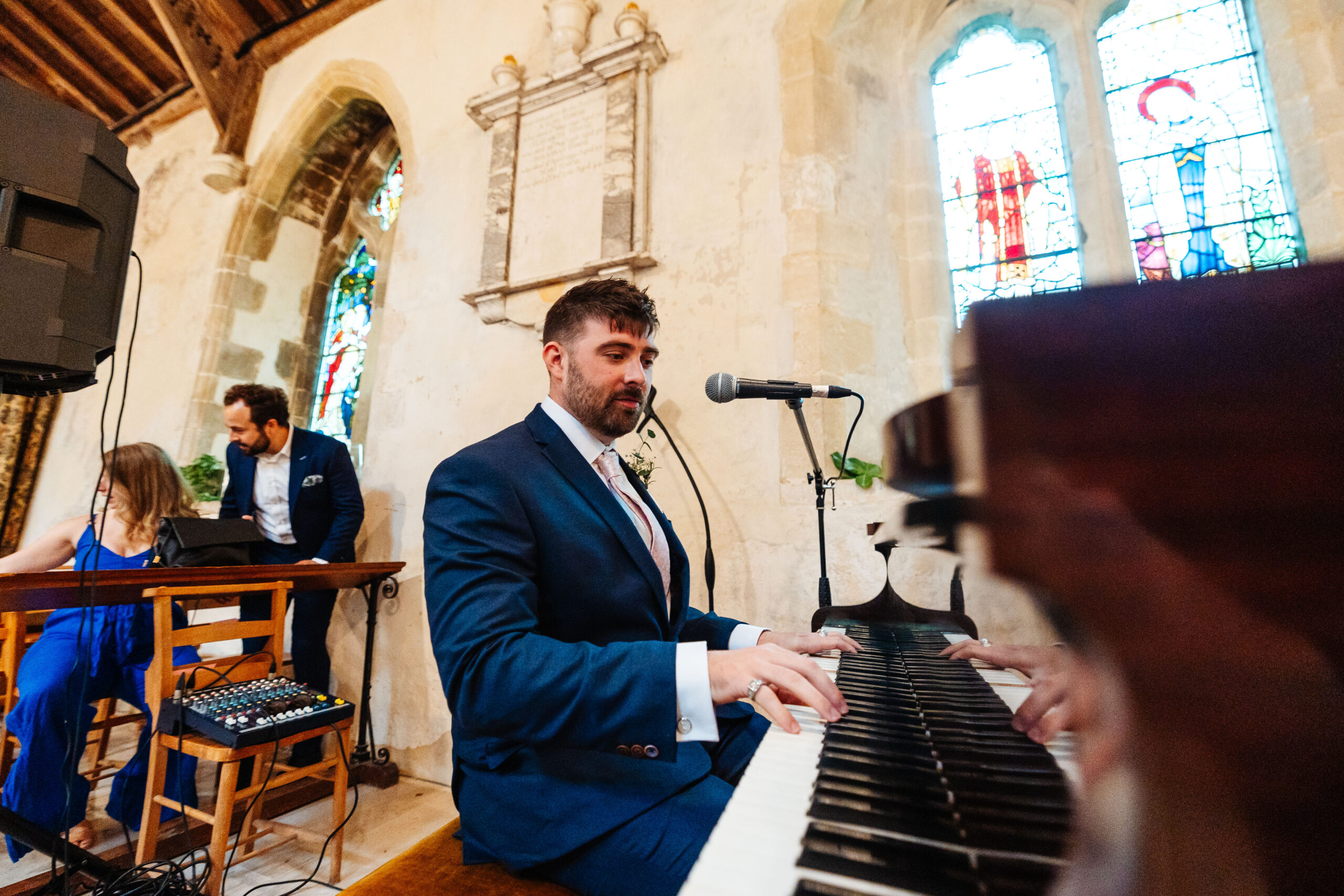 A man sitting down, playing the piano in a church. He also has a microphone near his mouth.