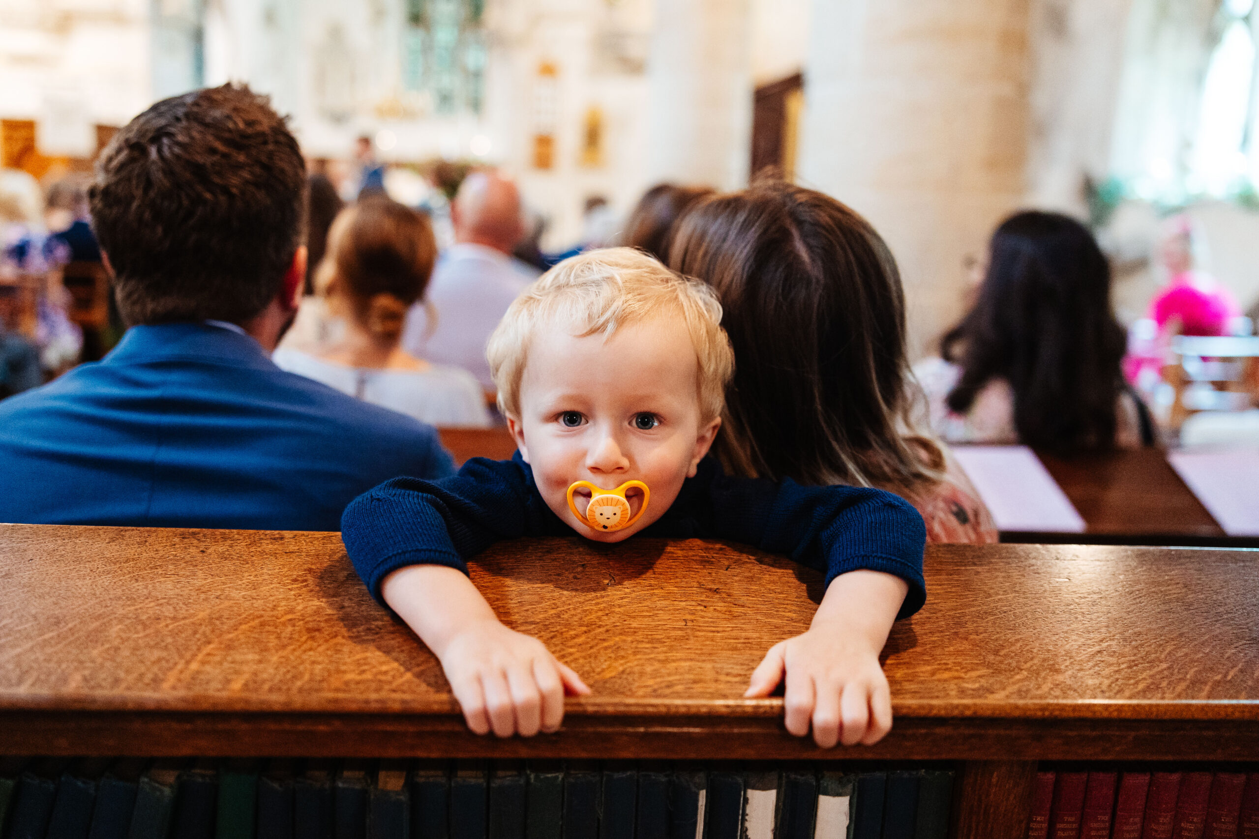 A baby boy looking at the camera and smiling with a dummy in. He is leaning over the pews in a church.