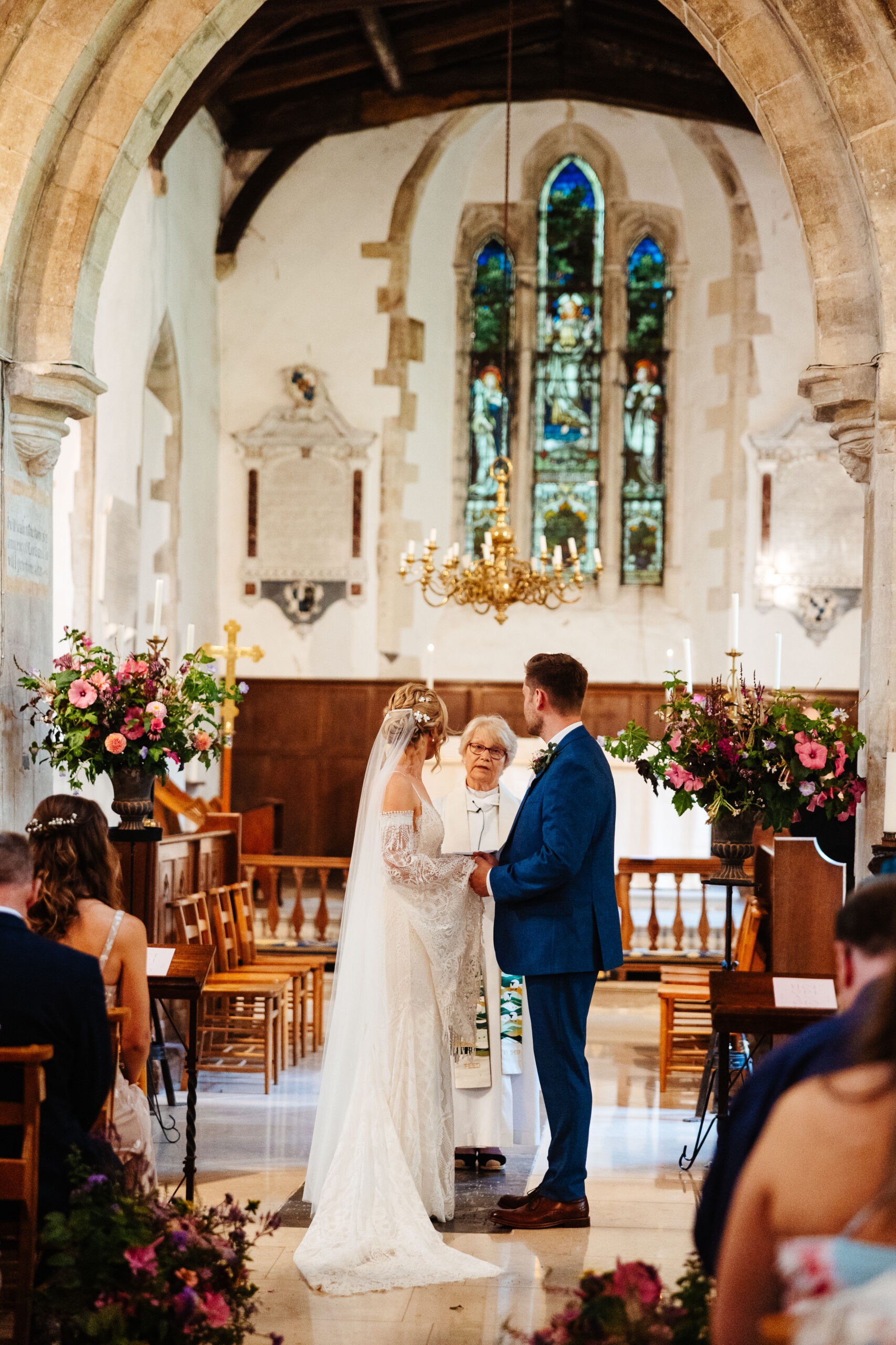 The bride and groom at the front of the alter in a church. You can see the stain glassed windows and a golden chandelier hanging from the ceiling.