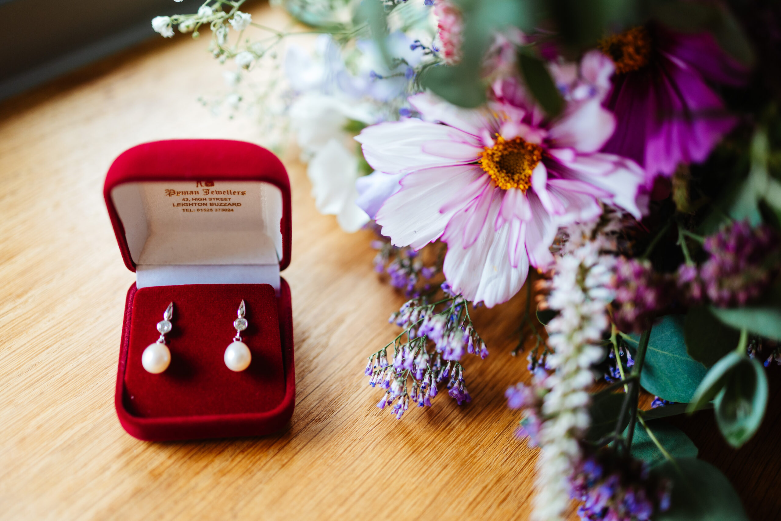 A pair of pearl earrings in a red, velvet box next to the bride's bouquet of flowers.