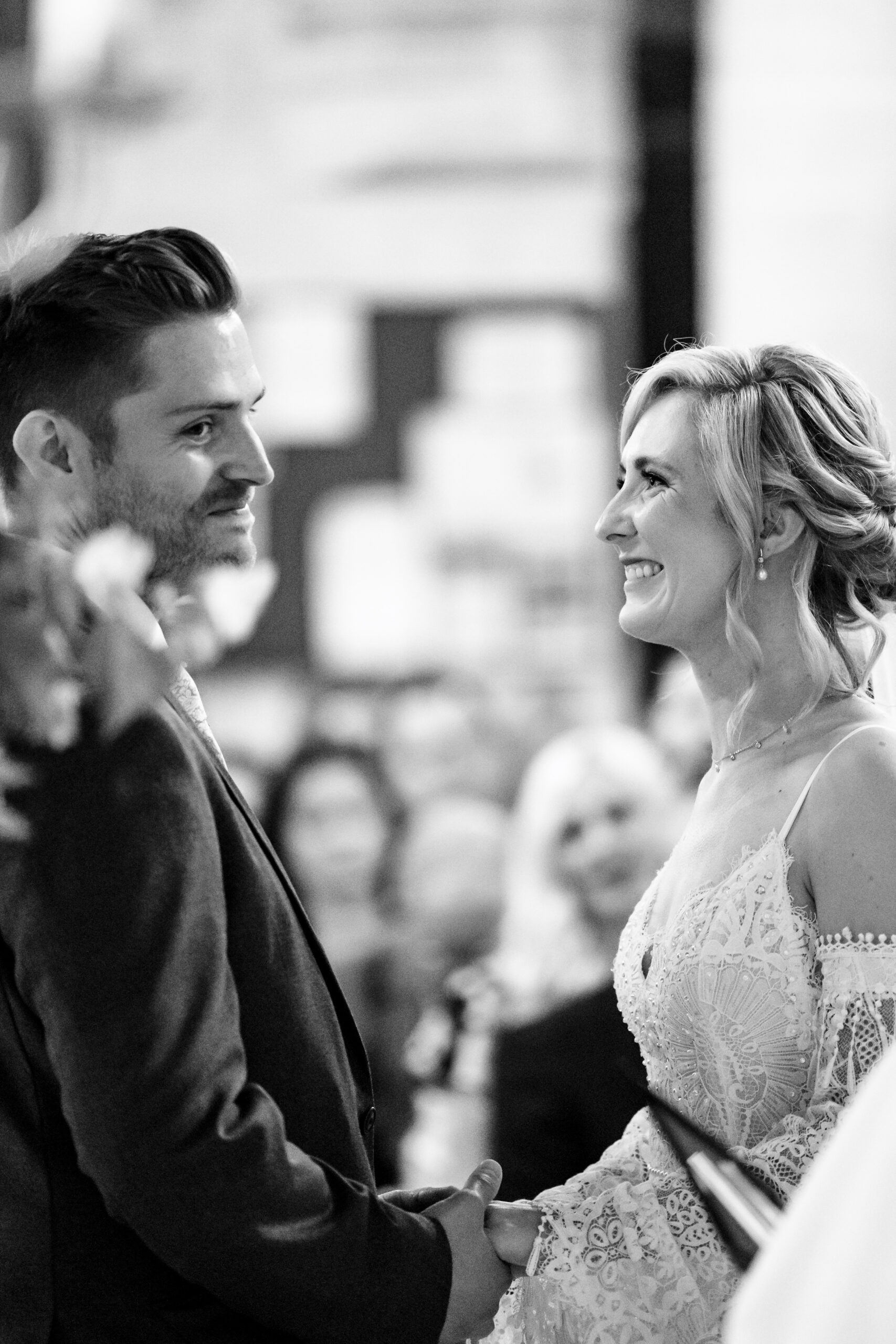 A black and white image of the bride and groom. She is smiling at the groom and he is smiling looking at the priest.