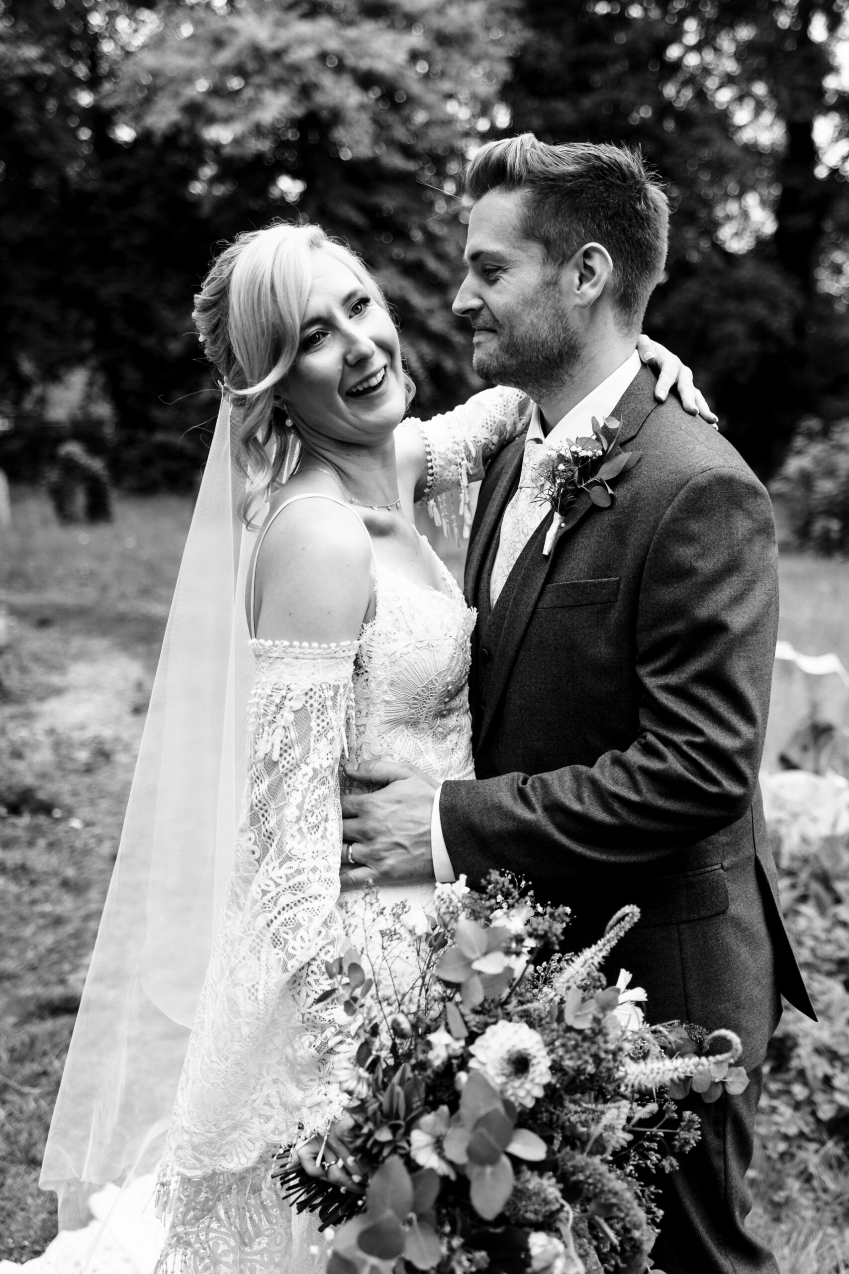 A black and white image of the newlyweds. The groom is looking at his bride and smiling. The bride is looking away.