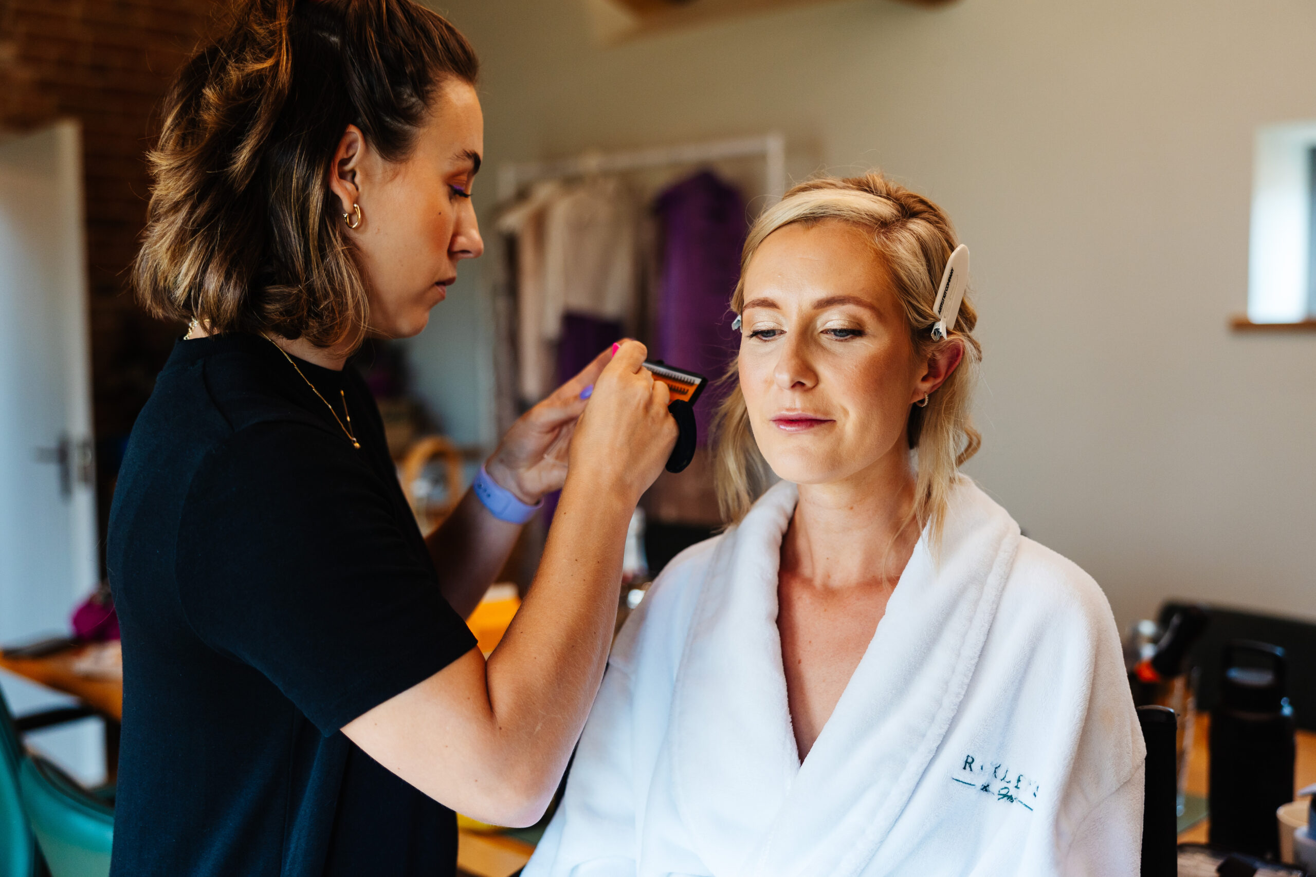 The bride having her make up done. She is in a white dressing gown and the make up artist is in a black t-shirt. She is applying eyelashes on to the brides eyes.