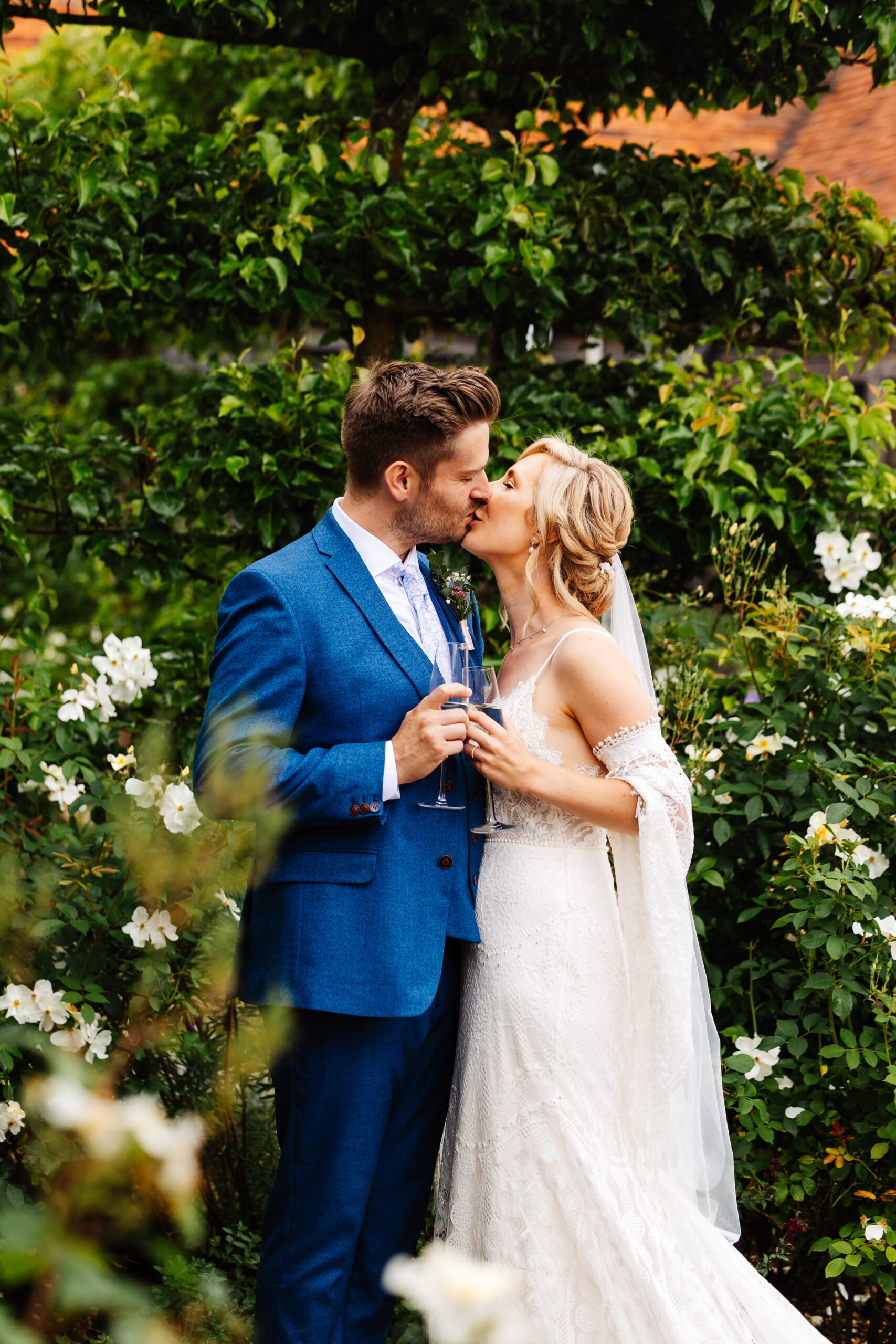 The bride and groom standing in front of some greenery outside. They are kissing and holding a glass of champagne in their hands.