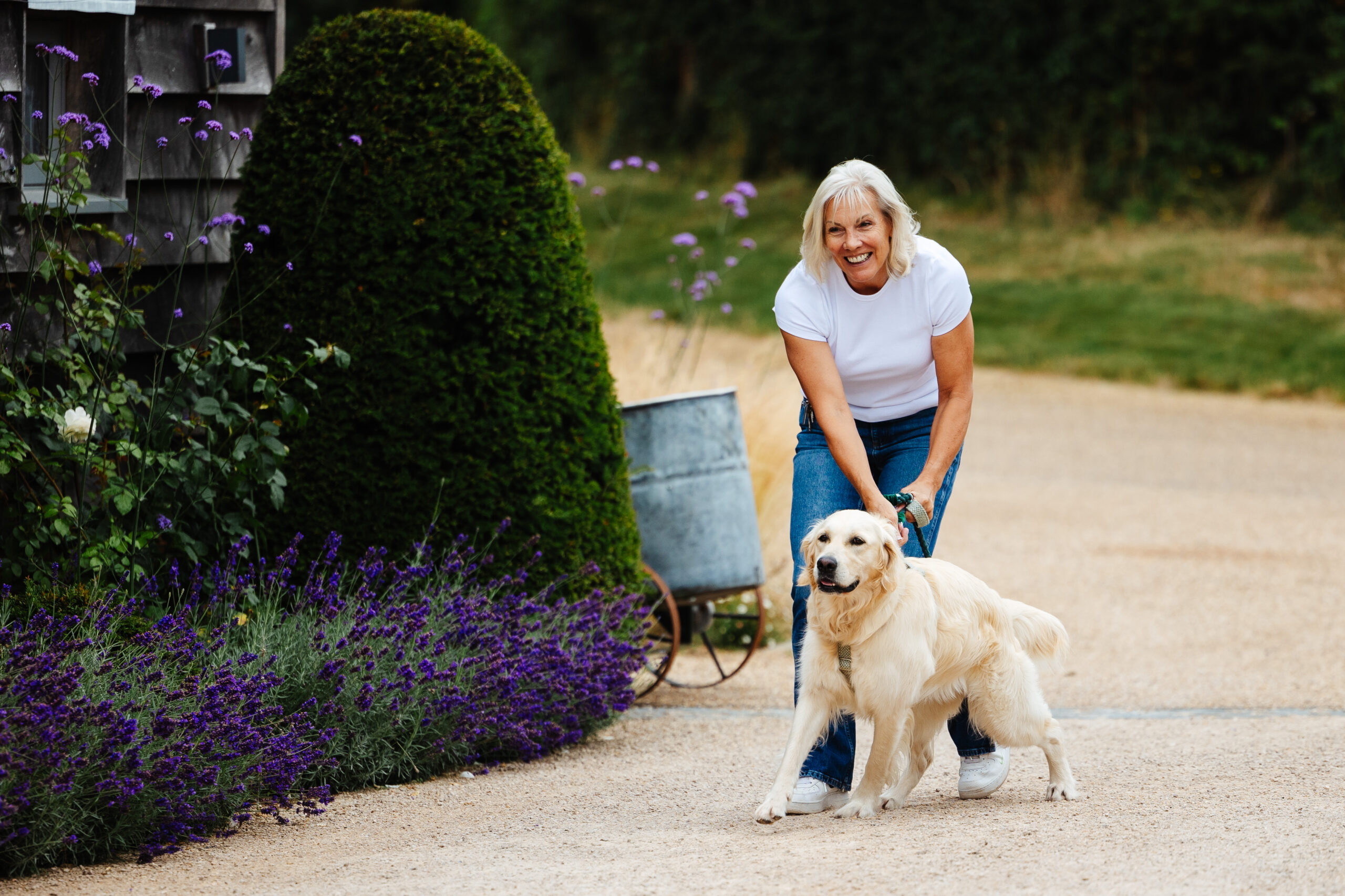 A lady in jeans and a white t-shirt. She is smiling and holding a golden retriever in her hands.