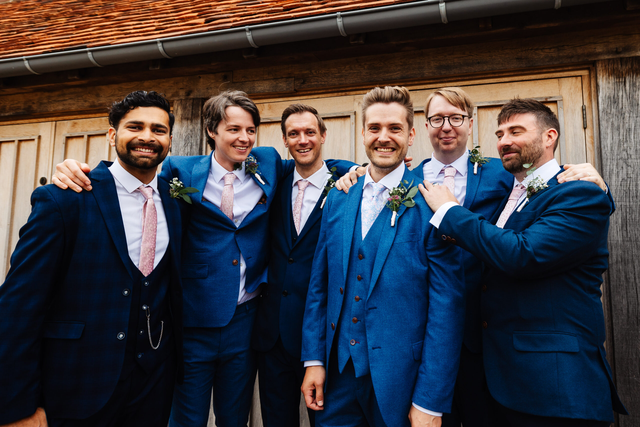 A group shot of the groomsmen. They are all in a variety of navy blue suits with pink ties on. They are surrounding the groom and look happy.