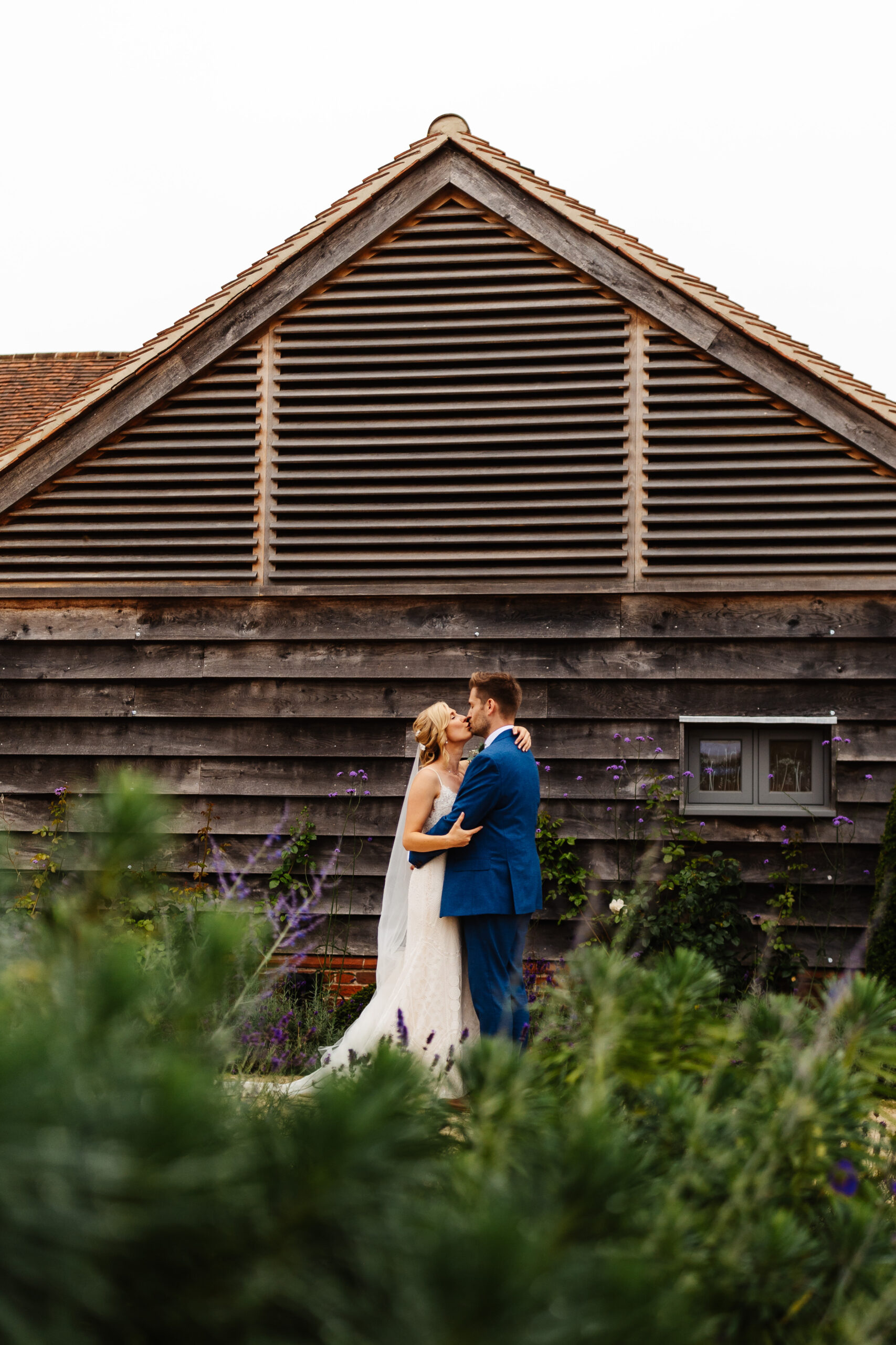 The bride and groom in front of a barn. There is greenery and flowers in front of the camera shot. They are kissing.