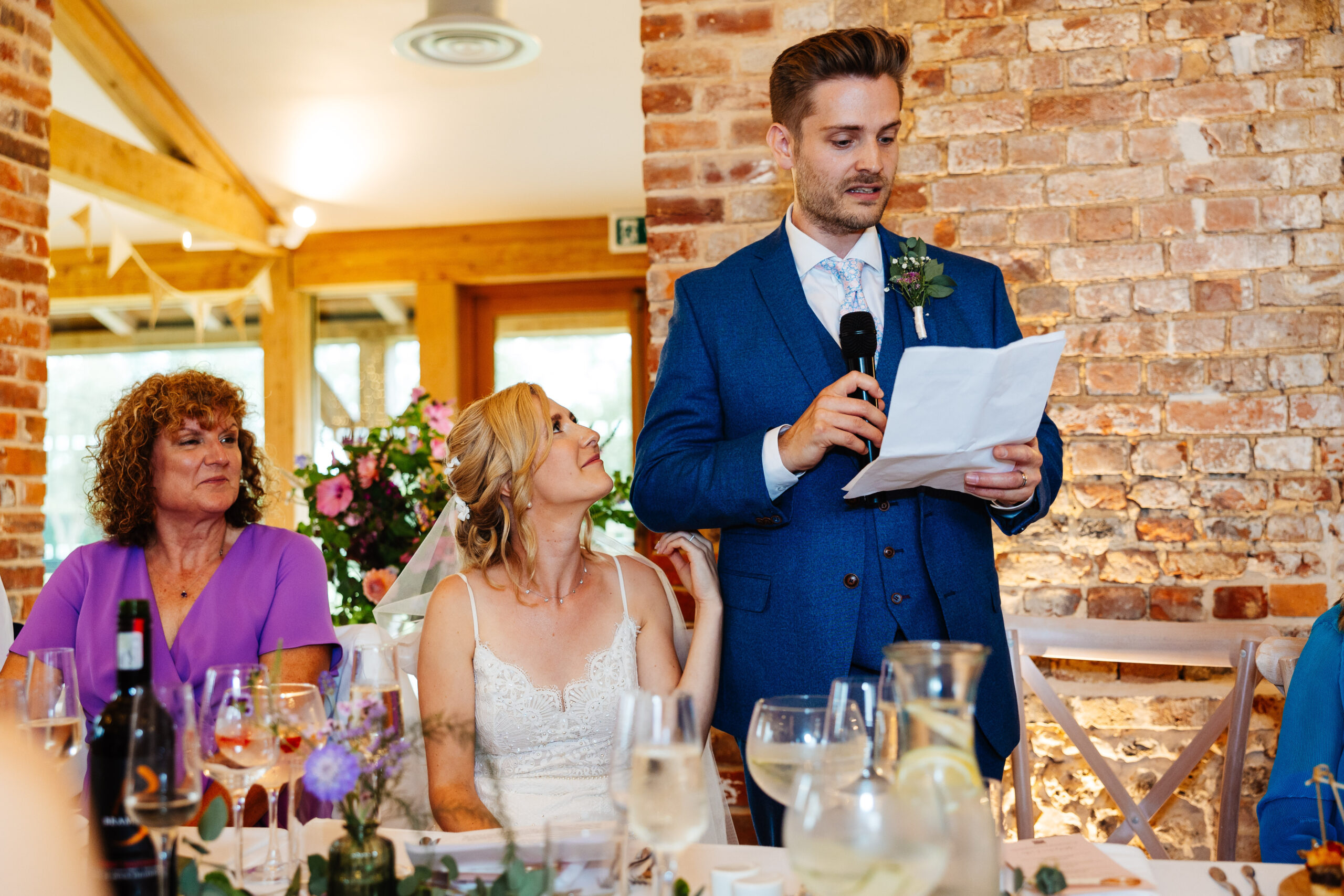 The groom standing up giving his speech. He is holding a microphone and his speech on printed paper. The bride is looking up at him and touching his arm with her hand.