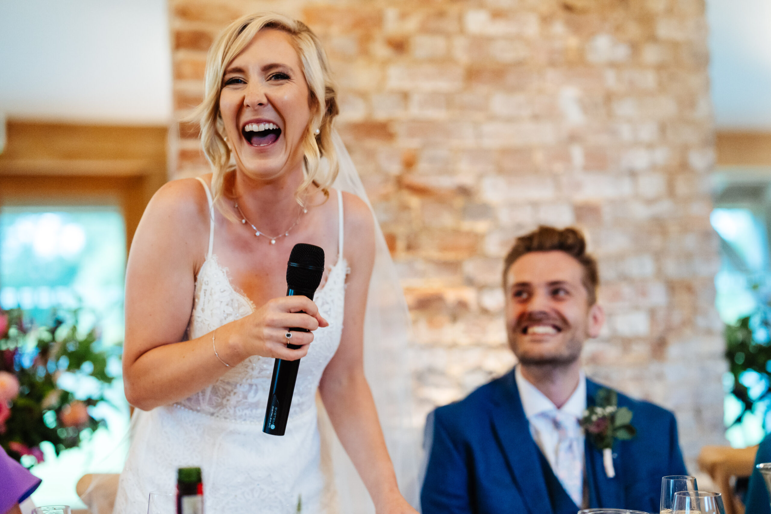 The bride giving a speech. She is holding a microphone and is smiling. Her groom is looking at her and smiling.