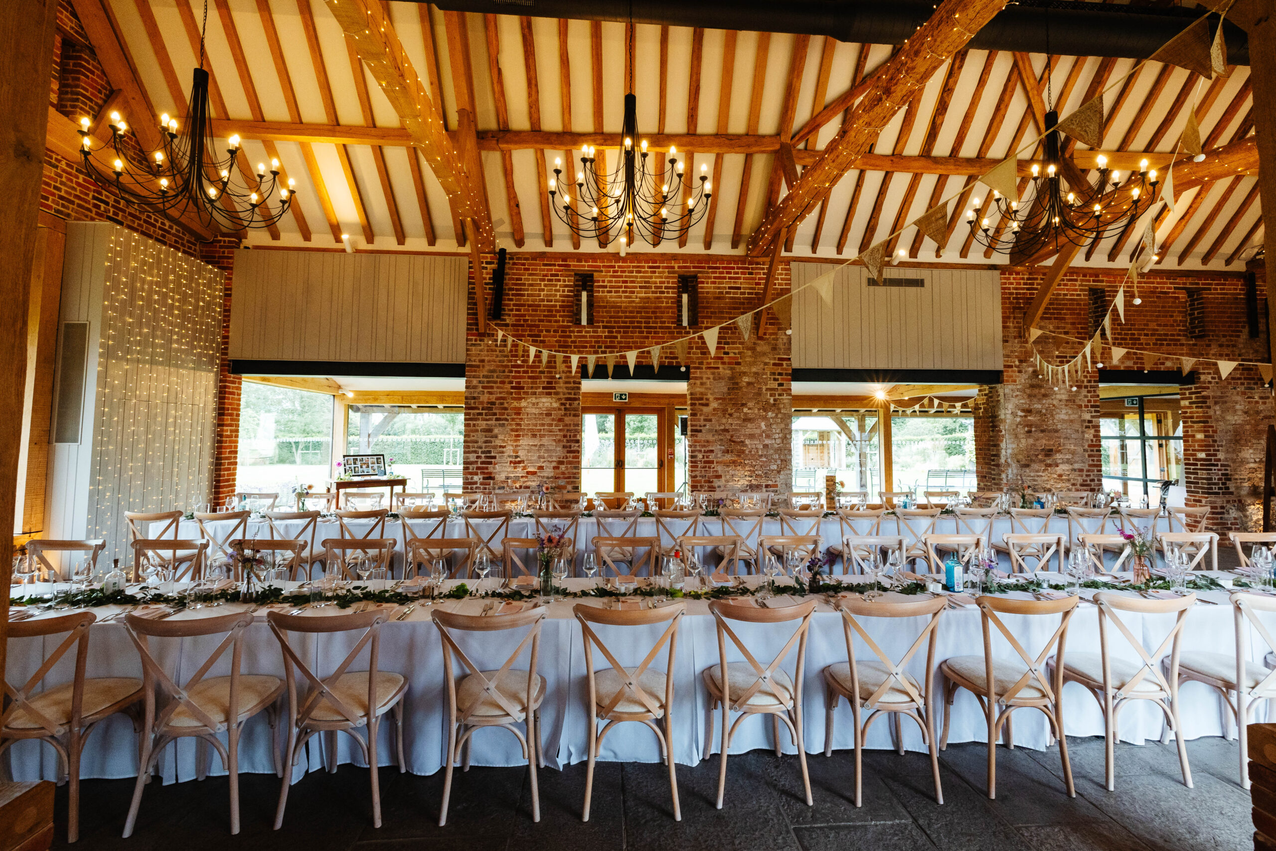 A barn dressed for a wedding. There are long tables with white table cloths, wooden chairs and bunting hanging from the ceilings.