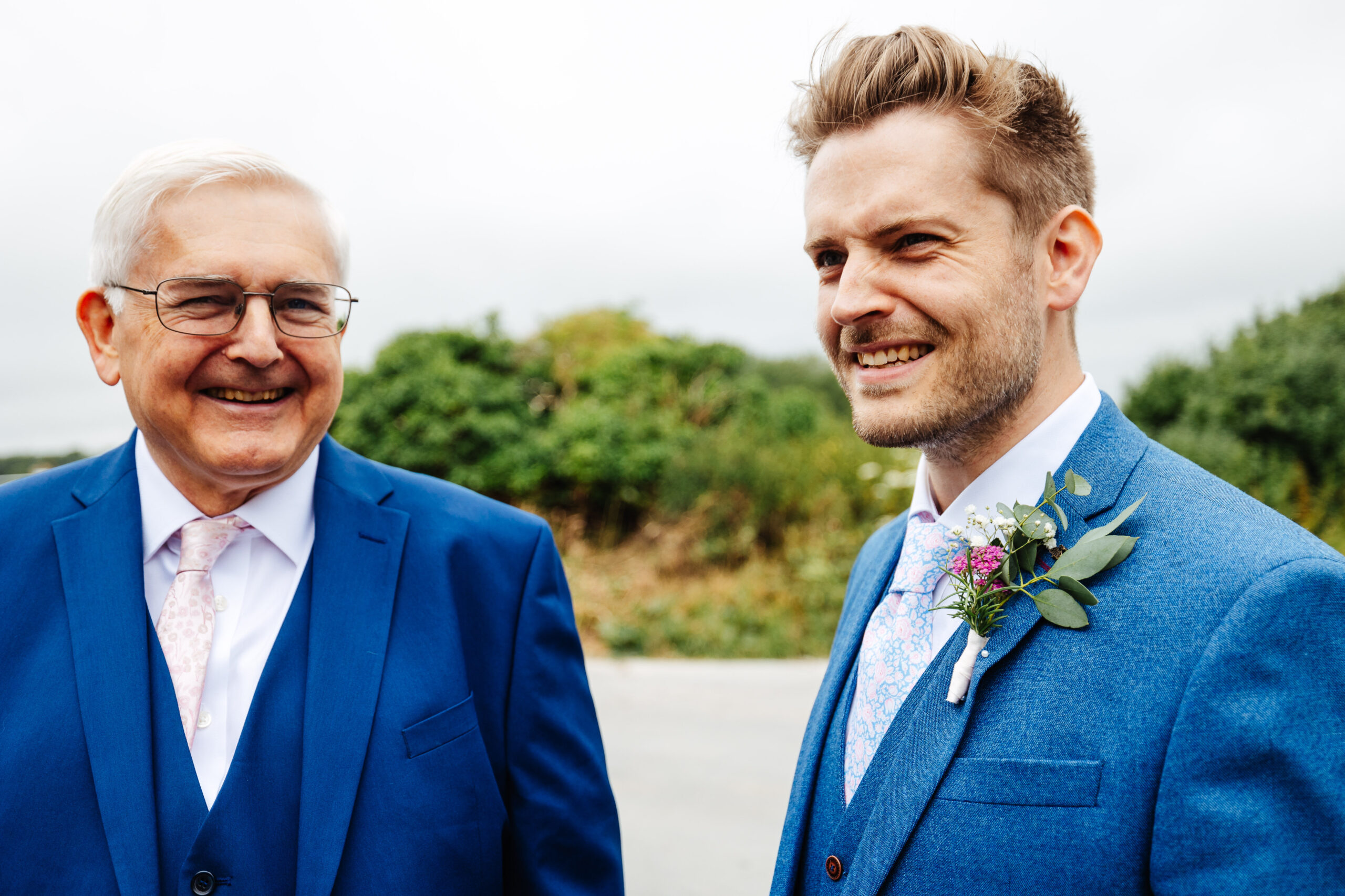 The groom and his father. They are both wearing navy blue suits and the groom has a small button hole of some flowers and some eucalyptus on his left shoulder.