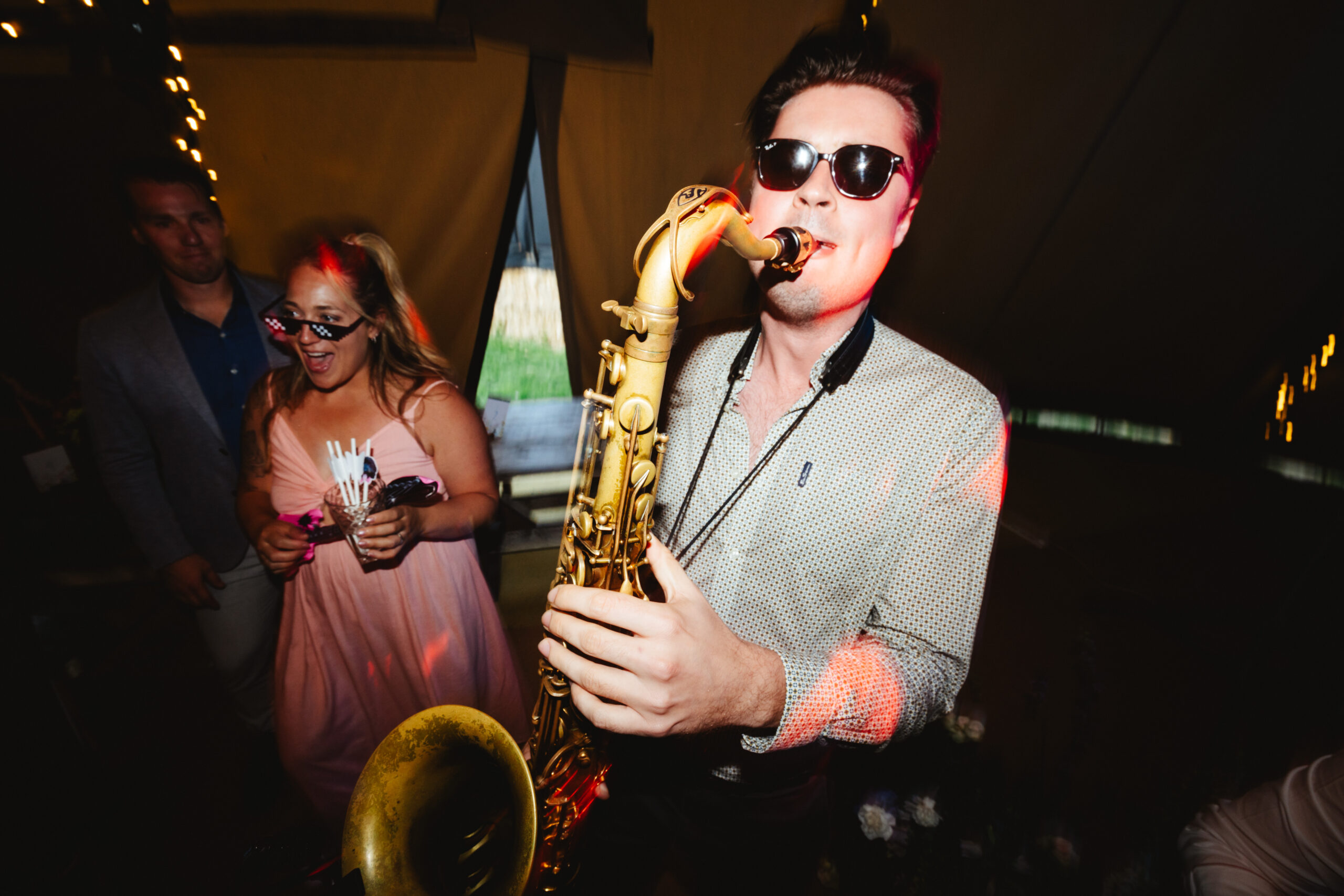 A saxophonist wearing sunglasses and is playing his saxophone in the tipi. People around him are dancing