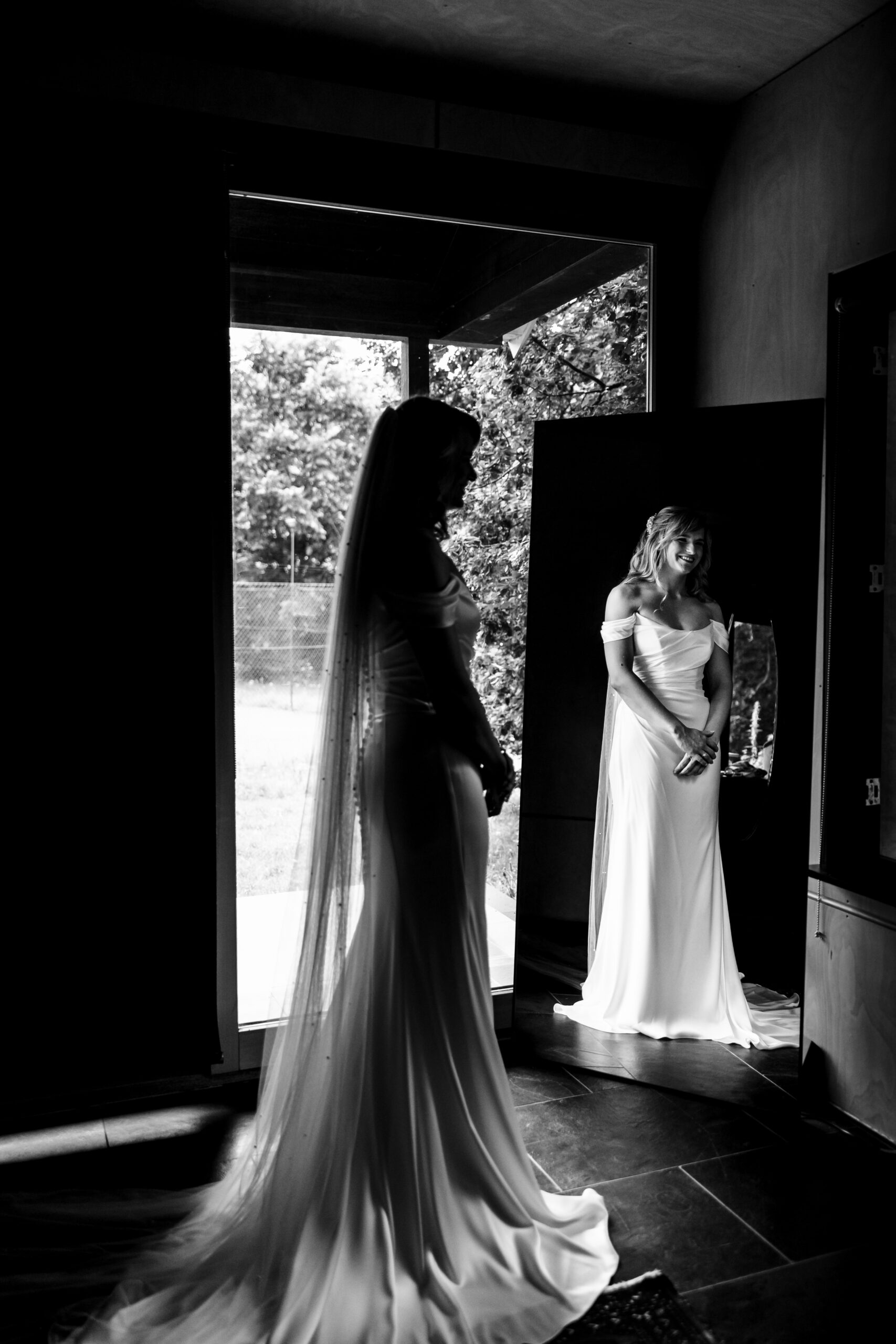 A black and white image of Olivia in her dress. It is long and flowing as is her veil. You can see Olivia both in the mirrored reflection of a long, free standing mirror as well as in the image itself.