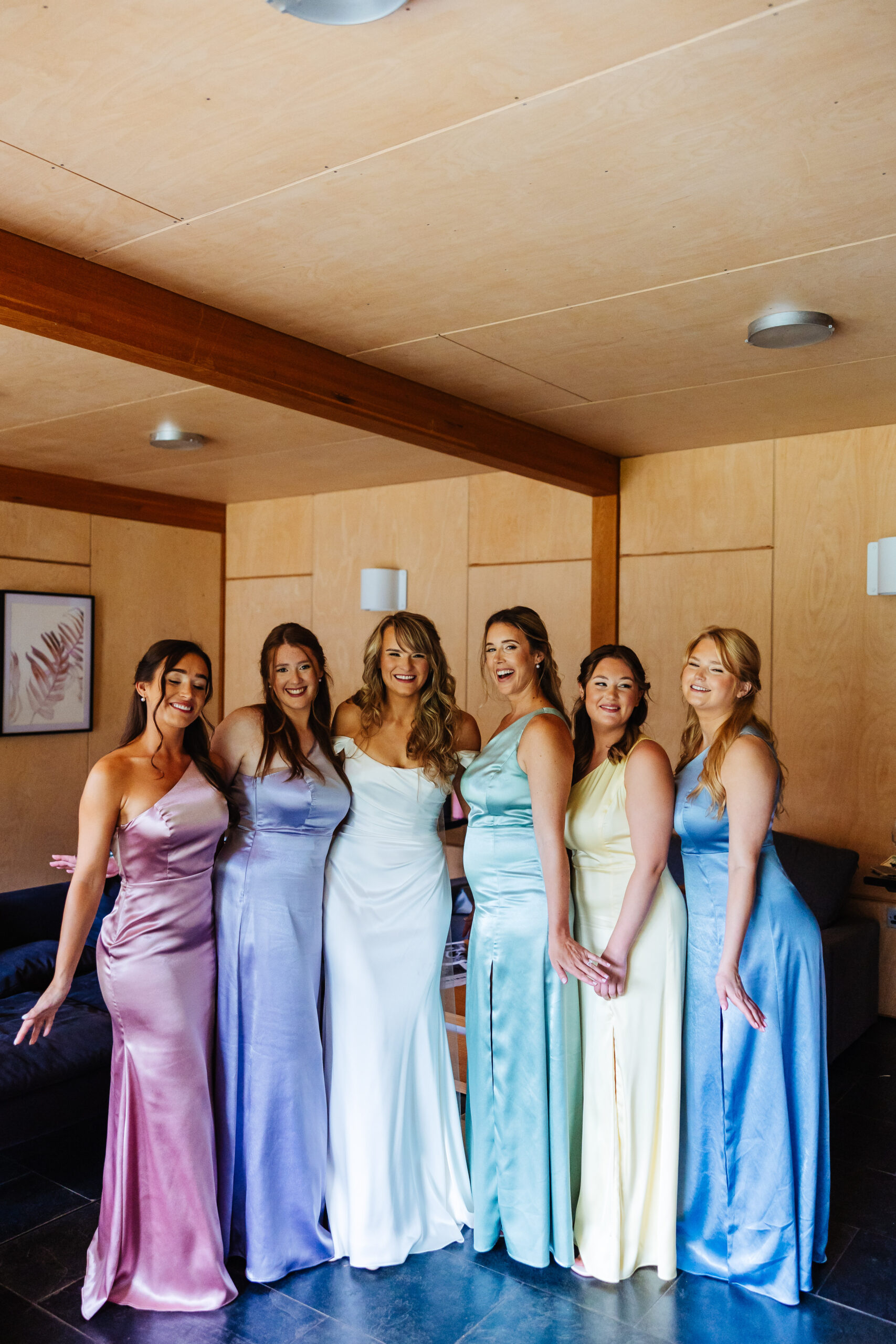 The bride in an off-the-shoulder white wedding dress and her bridesmaids who are wearing pastel coloured dresses; pink, purple, green, yellow and blue.