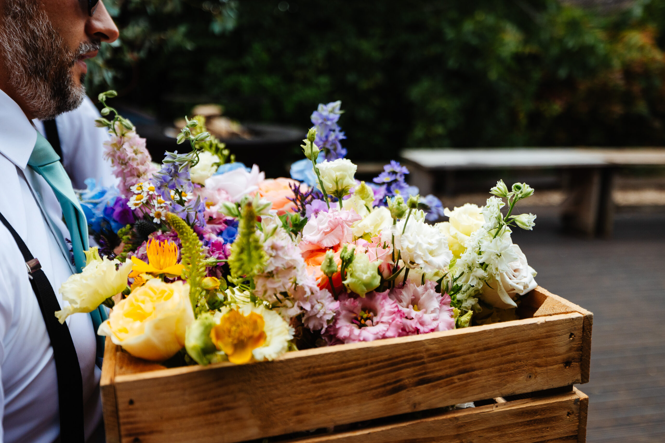 A man carrying a pallet box of flowers. The flowers are a mixture of pink, yellow, blue, purple and white.