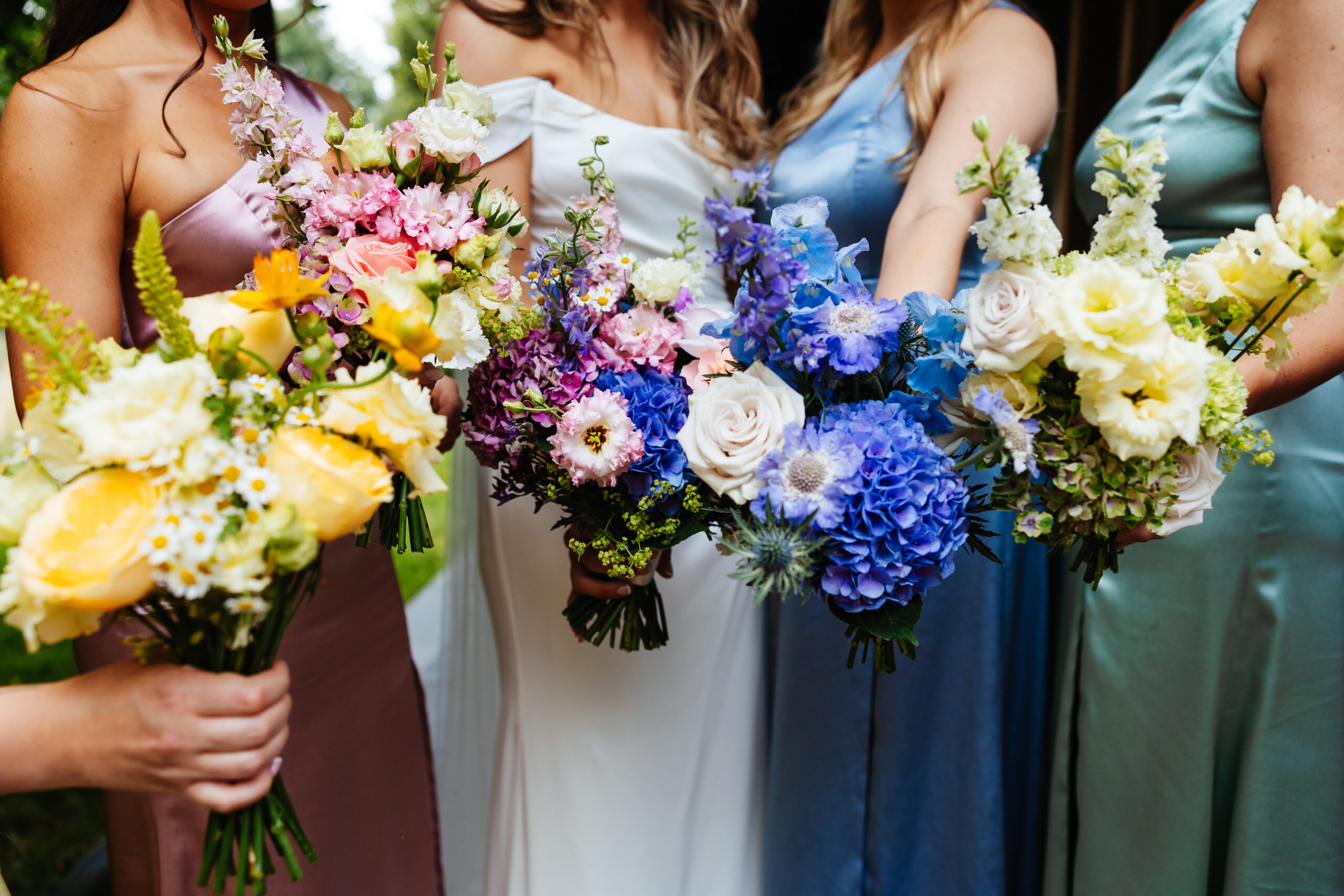 Each bridesmaid is holding a bunch of flowers that matches their coloured dress; yellow, pink, purple, blue and yellow.