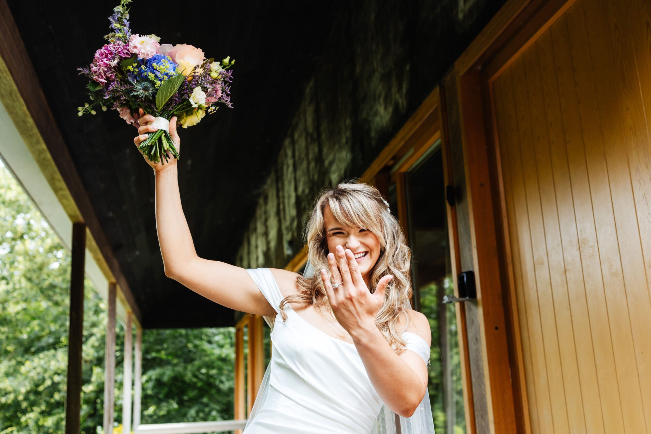 The bride in her wedding dress, outside, holding a bouquet of flowers in one hand up high and showing off her engagement ring on her other hand.