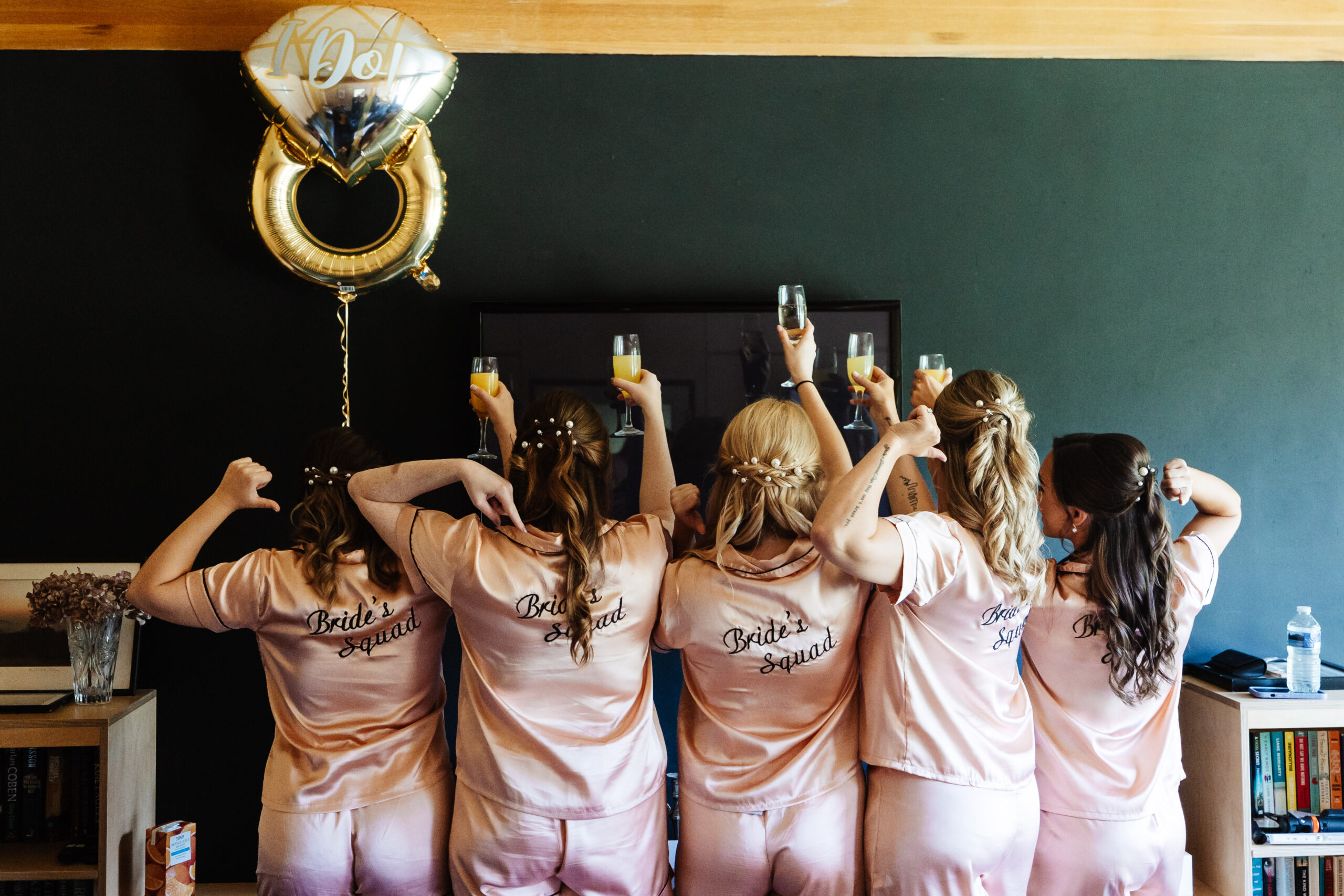 The bridesmaids with their backs to the camera. They are in pink, silk pyjamas with Bride's Squad on the back and they are holding a glass of prosecco up in cheers.