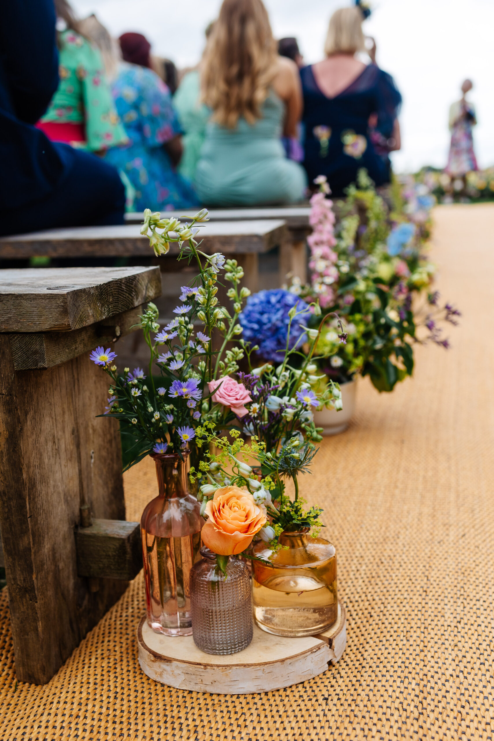 A photo down the aisle; a wooden log with small vases of flowers on it containing flowers in purple, orange and pink.