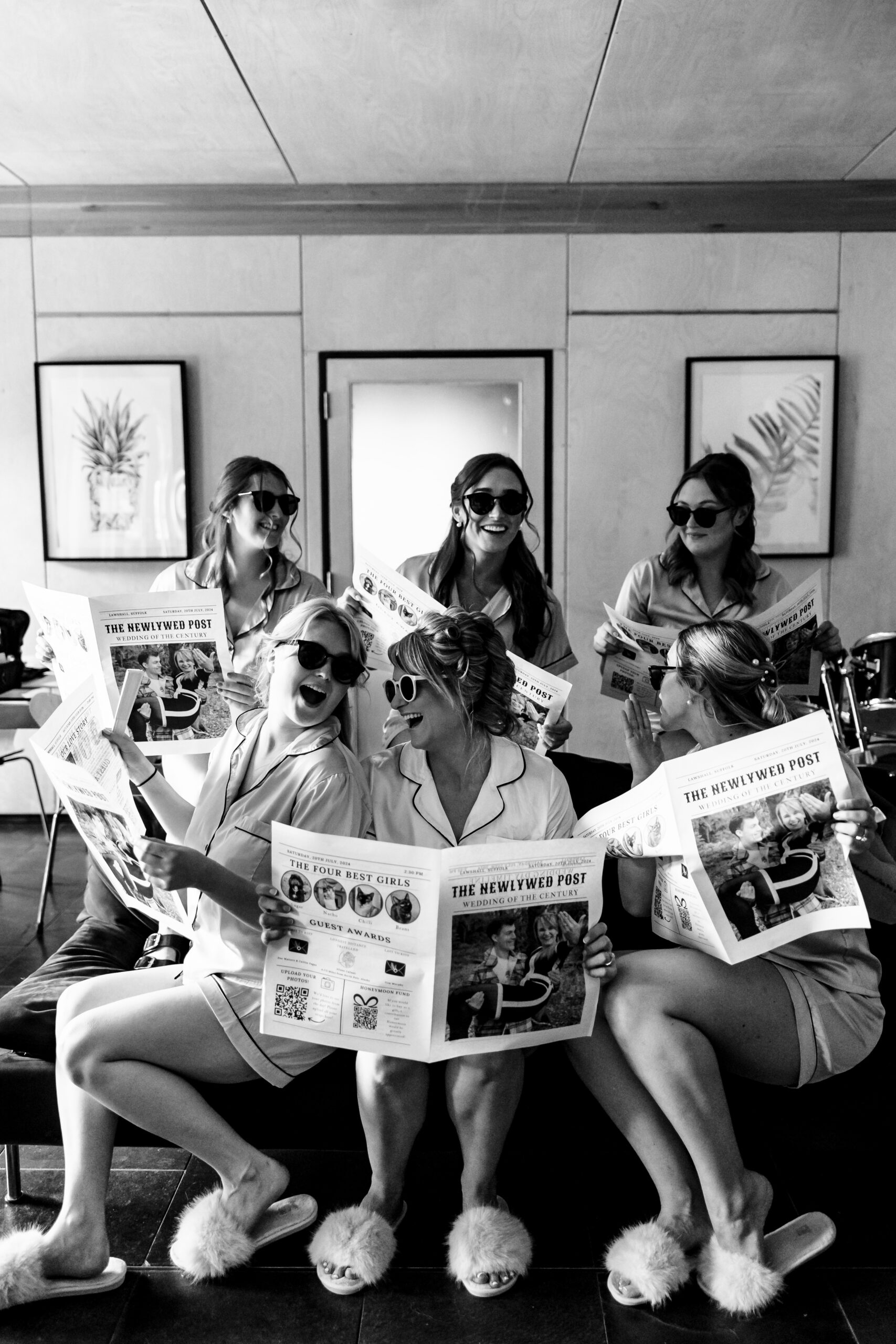 A black and white image of the bridal party holding newspapers featuring the bride and groom on the front page. They are laughing and all wearing sunglasses indoors. They are having fun.