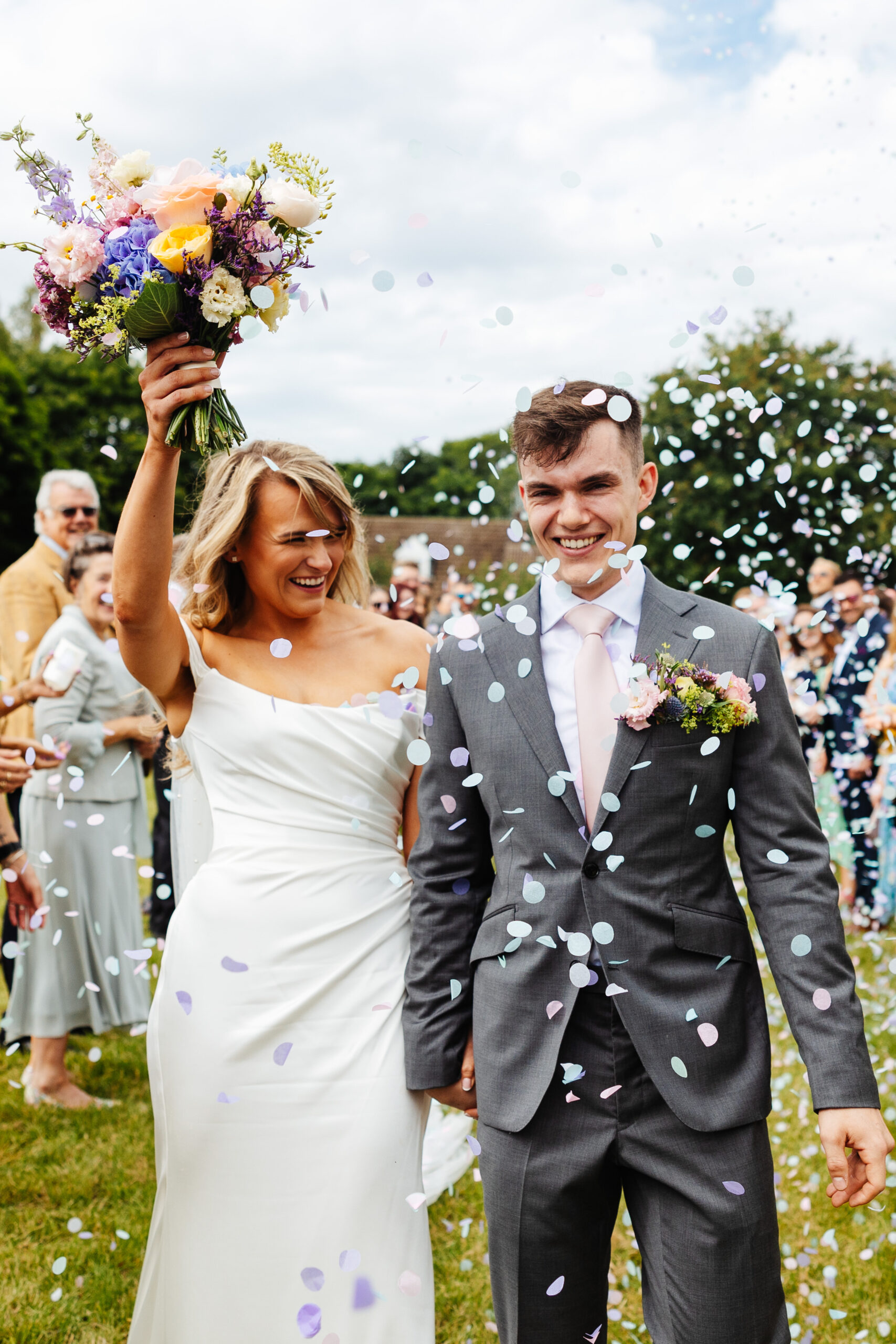 The bride and groom walking through confetti. It is surrounded around them. They are smiling and happy and the bride is lifting her arm in the air with her flowers in her hand. 