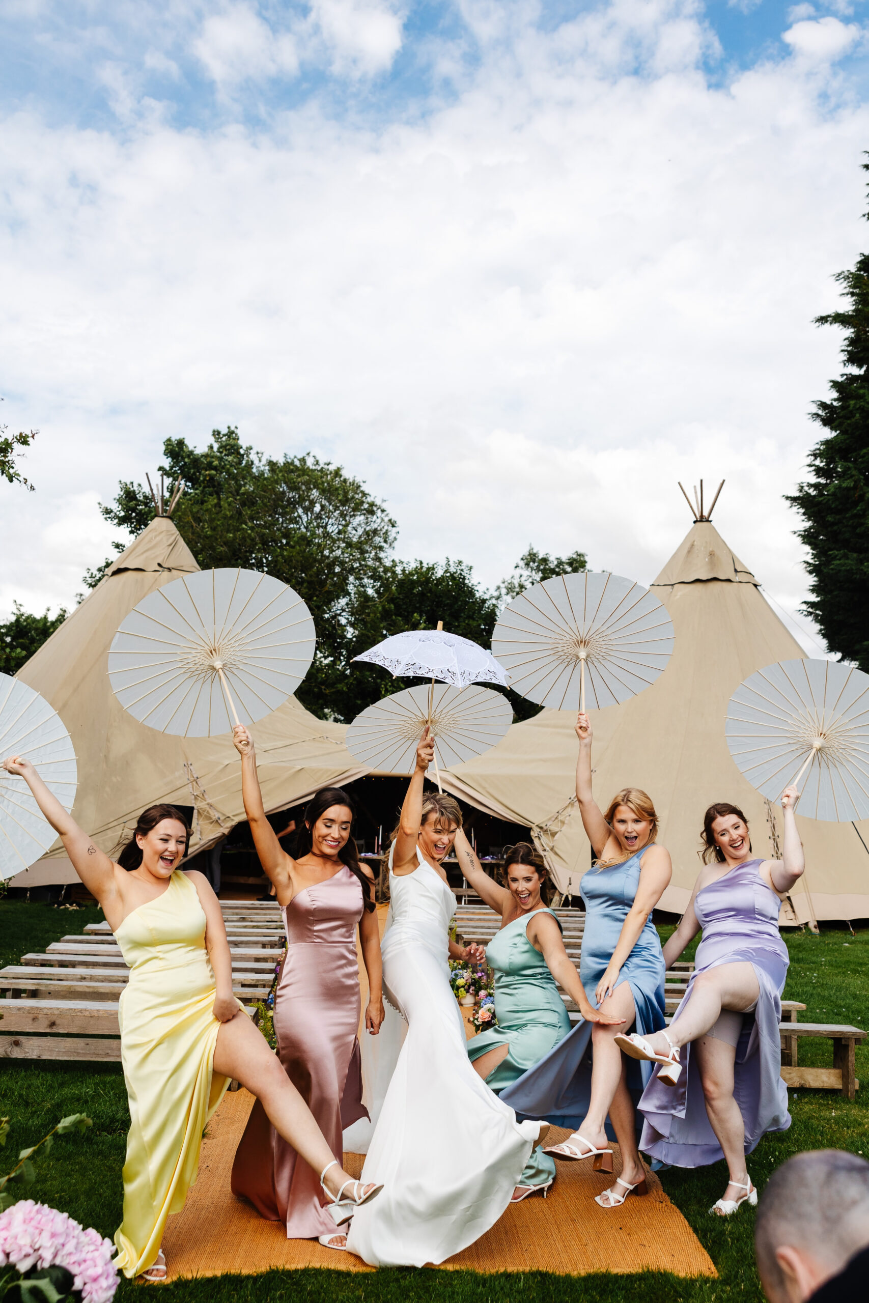 The bridesmaids and bride having fun! They have white umbrellas in the air and they are kicking one leg in the air. They are all smiling.