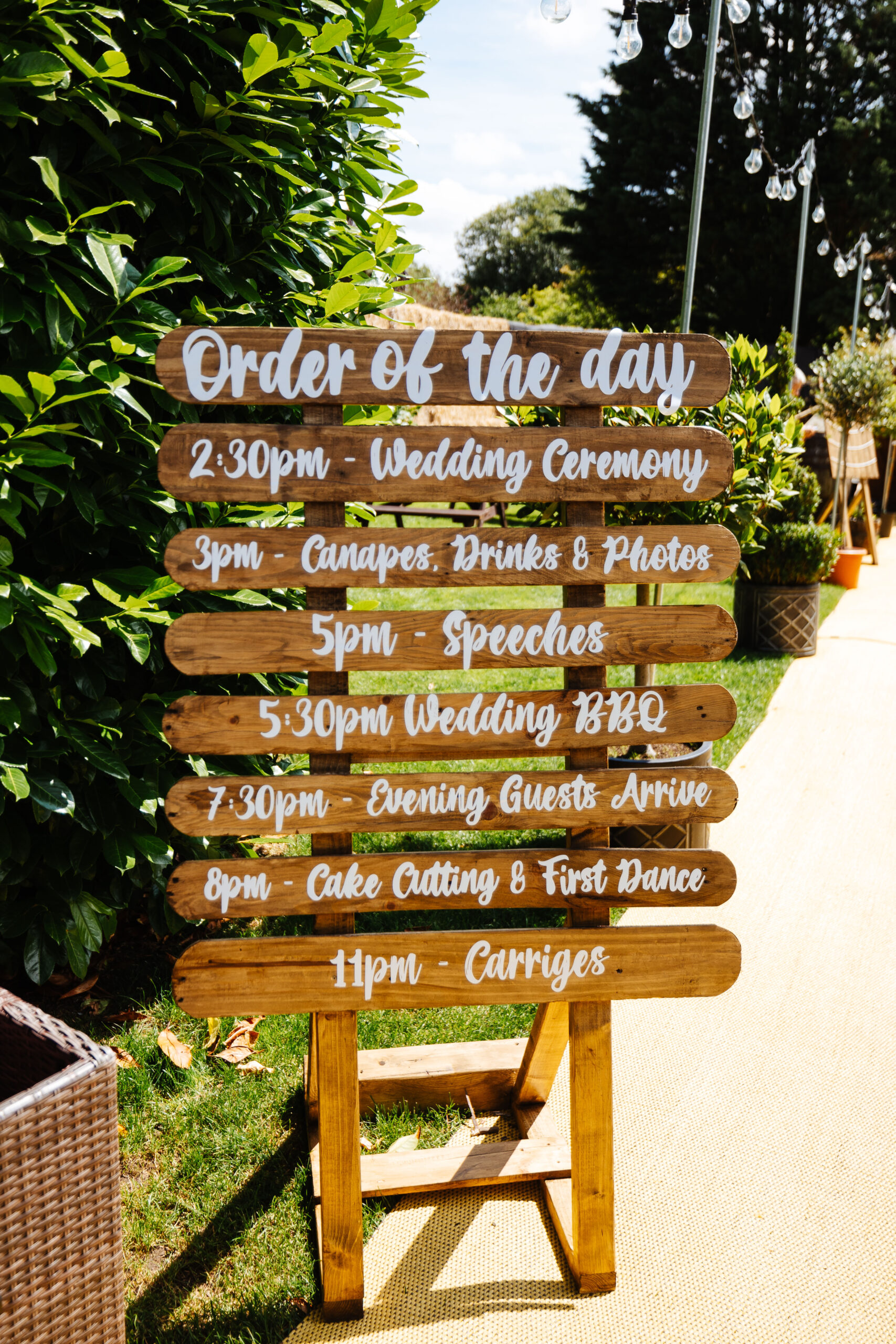 A photo showing the order of the day. It is outside and you can see bright green trees and bushes behind it. The order of the day is written in cursive, white writing.